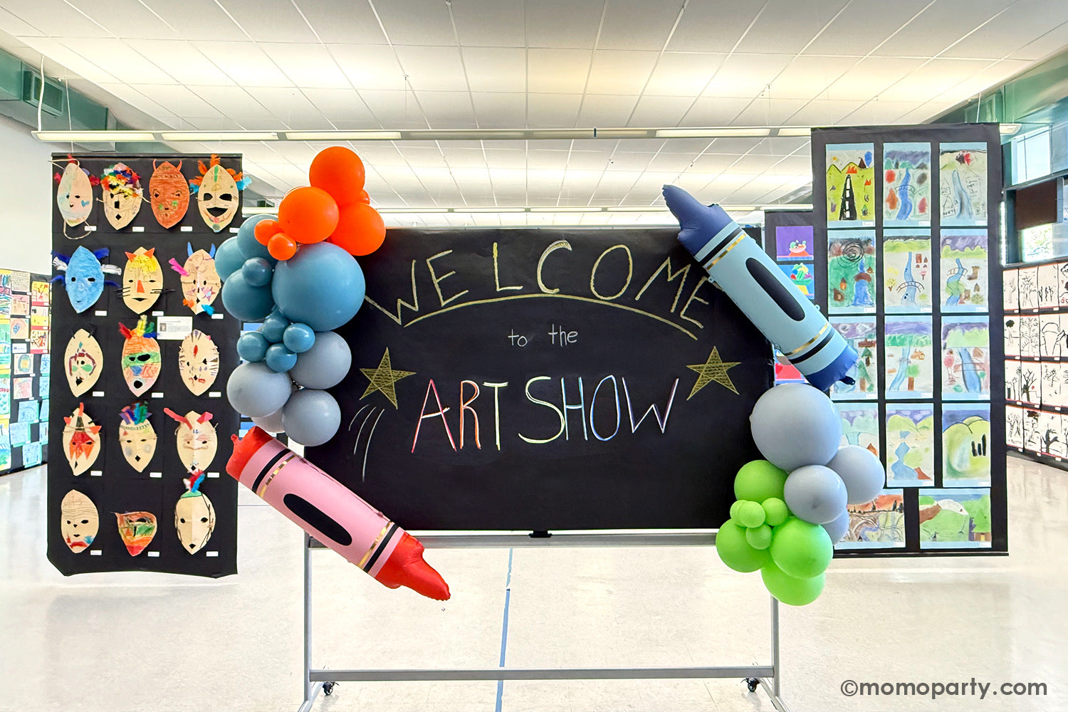 A school chalkboard decorated with a festive and colorful balloon garland along with Momopartys's 40" x 9" blue & red crayon shaped foil balloons at a kid's art show with arrays of kid's artworks in a school event. This easy balloon installation with bright color and fun design is also perfect for kid's school themed celebration, be it a back to school party, a first day of school celebration or any kids event at school.