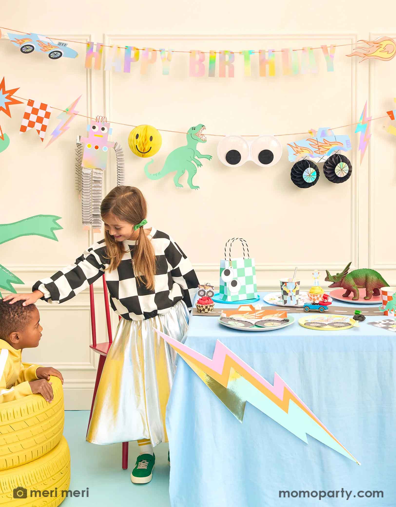A fun themed kid's birthday party decorating set up featuring Momopartyss Momopartys's Toybox Party Collection by Meri Meri. With the retro inspired and checker board designs,  featuring elements from kid's toy boxes including dinosaurs, monster trucks, race cars, googly eyes. robots and lightning bolts, these party supplies including a large party garland on the way with HAPPY BIRTHDAY message, and a table of fun and festive party tableware. Two kids playing next to the party table having a good time.
