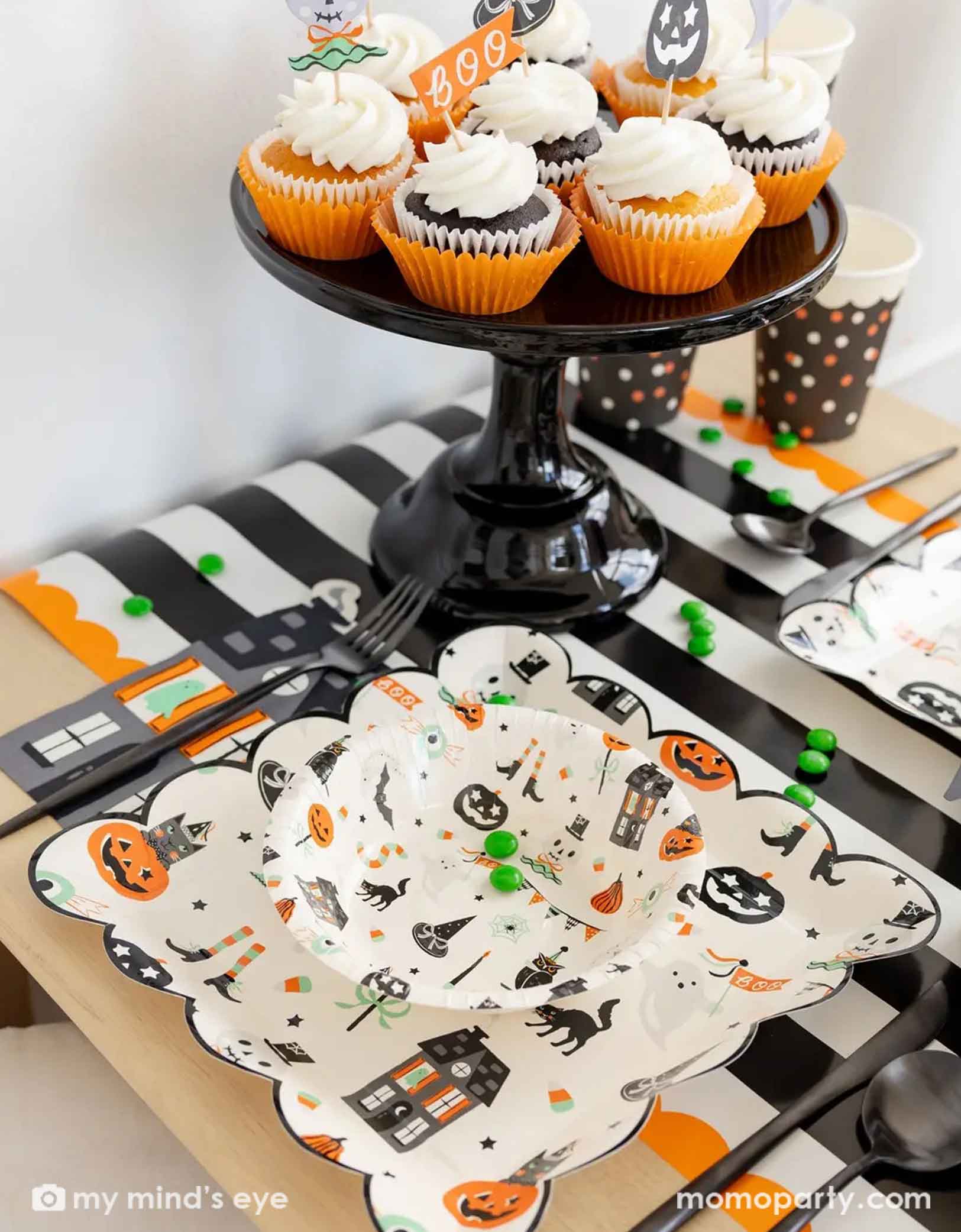 A side shot of a festive Halloween-themed table setting with Momopartys's Trick or Treat dinner plates and bowls, the haunted mansion shaped napkins and black cutlery set on a black orange and white striped table runner fro Momopartys. On the table there's a cauldron filled with green gum balls and some green candies spread across the table. A few cupcakes topped with Halloween themed toppers on a black cake stand next to it. A perfect set up for kid's friendly Halloween celebration.