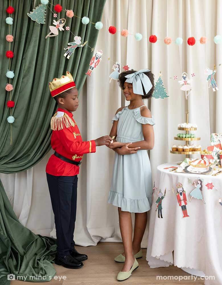 A girl and a boy dressed as the Nutcracker characters in a Nutcracker themed holiday party. They're standing next to a party table filled with Holiday treats and Nutcracker themed party supplies. In their back there's a stage curtain in satin green and beige which is decorated with Momopartys's Nutcracker 6' garland with pom-poms which adds a festive touch to the Holiday decoration.