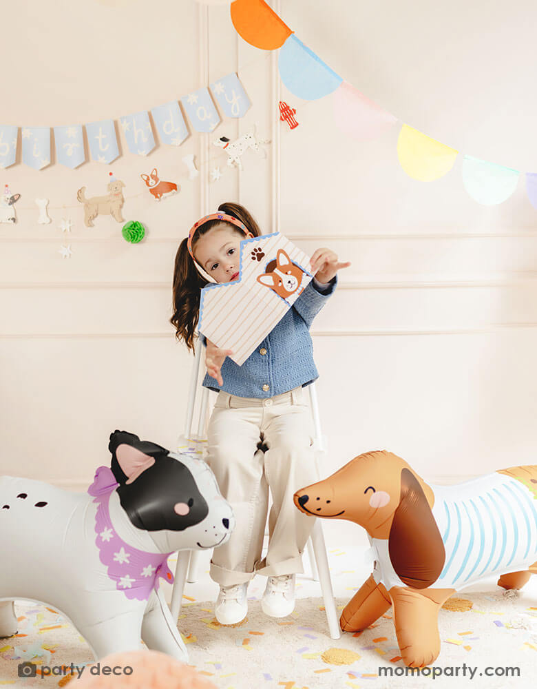 A school aged girl holding Momopartys's dog house shaped party box standing in between two dog shaped standing foil balloon in a puppy themed birthday party. On the wall there's a dog themed birthday party banner and a multi-colored bunting garland, making this a festive and fun scene for kid's birthday celebrations.