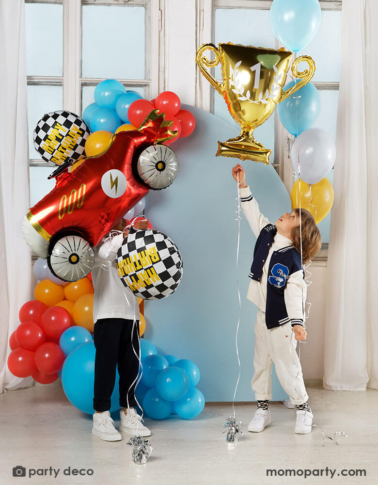 Two boys playing with Momopartys's race car themed foil balloons including a red vintage race car shaped balloon, two checkered flag happy birthday foil balloons and a gold trophy shaped foil balloon, both kids standing in front of a pastel blue party backdrop wall which is decorated with colorful balloon garland with race car inspired colors including red, light blue, orange, gray and yellow. All makes this a festive and fun kid's birthday celebration.