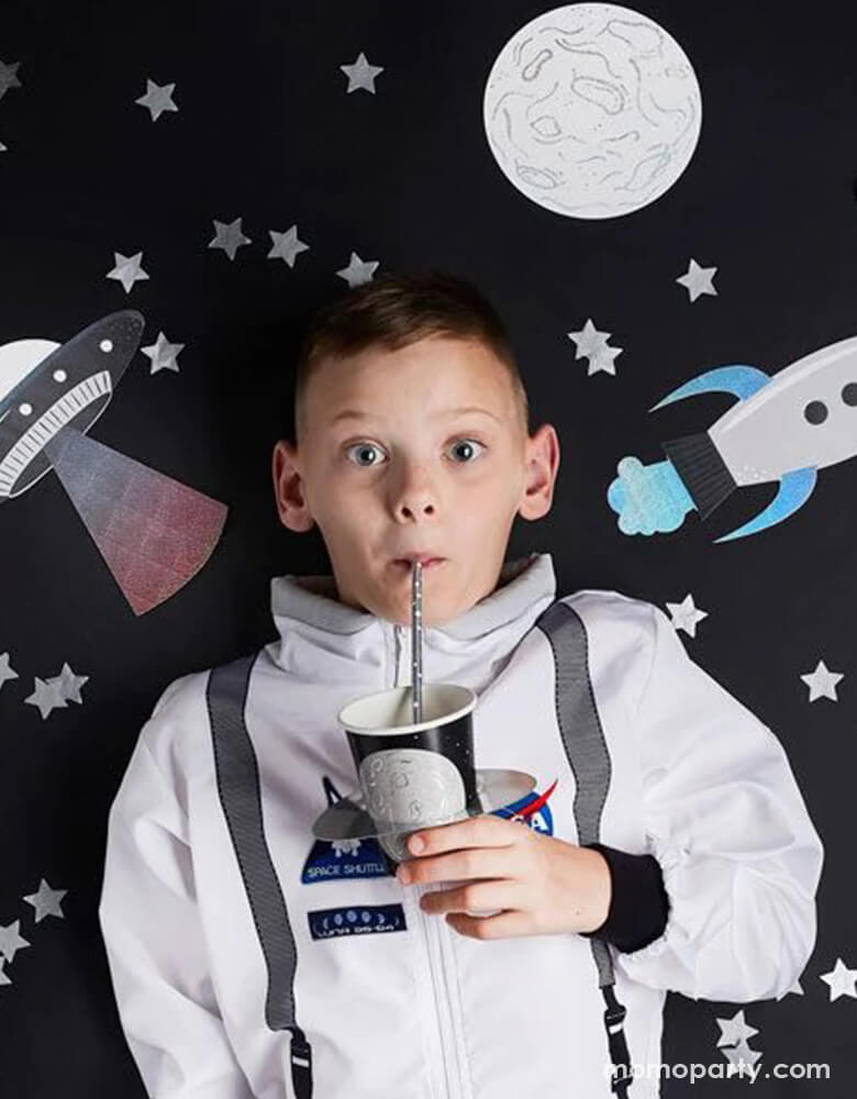 A boy in his astronaut suit holding Momopartys's 9" planet paper cups by Hooty Balloo. He's drinking from the cup with a silver foil straw, in his back the wall is decorated with space themed cutouts including rocket ships, UFOs, moon, and stars.