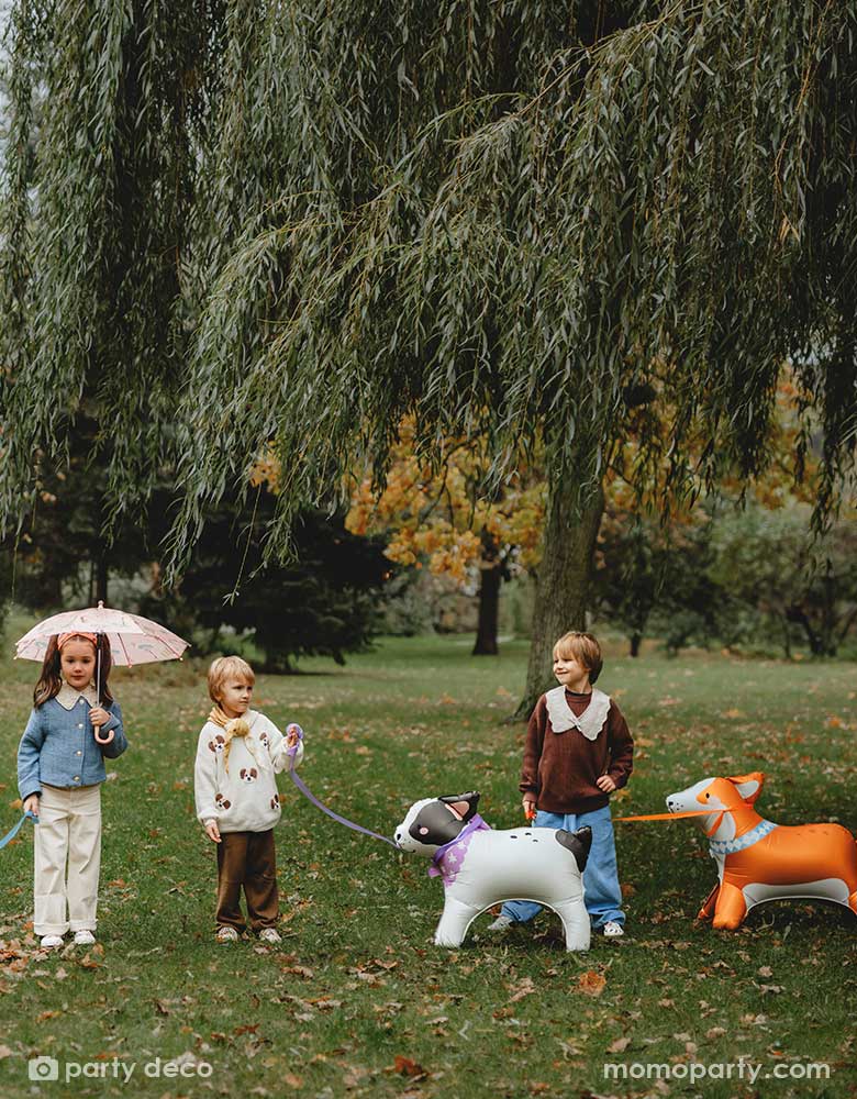 Three kids playing pretend play walking Momopartys's bull dog and Corgi standing foil balloons in a park. The girl holds an umbrela and the boys are talking. 