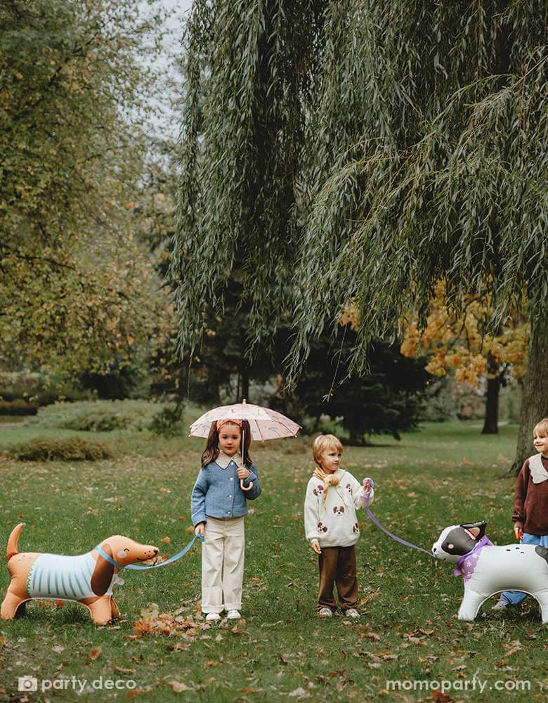Three kids playing pretend play walking Momopartys's French bull dog and Dachshund standing foil balloons in a park. The girl holds an umbrella and the boys are talking to each other having fun in the park.
