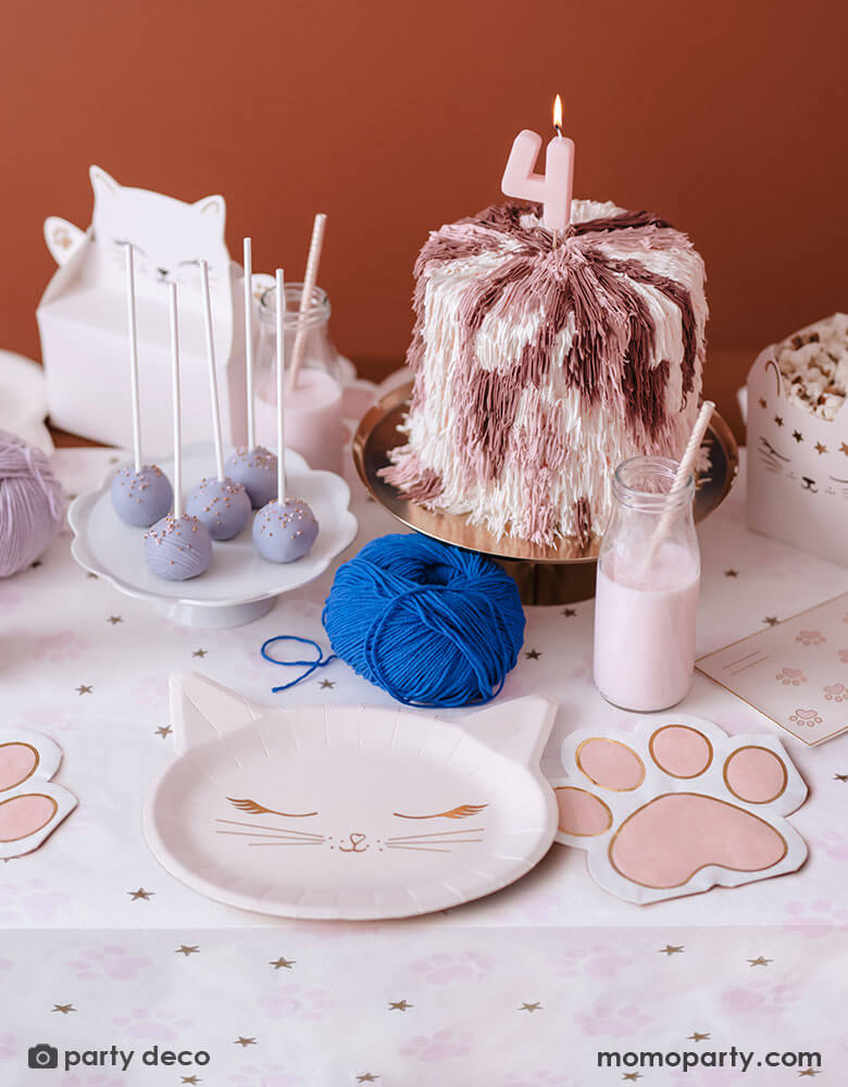 A cat themed party table featuring various cat themed party supplies from Momopartys including party plates, pink paw shaped napkins, cat shaped treat boxes filled with popcorn, small milk bottles with pale pink drink and a table of treats, snacks and a cake topped with a pink number 4 candle in pastel pink and lilac colors, making this an adorable inspo for kid's kitty cat themed birthday celebration.