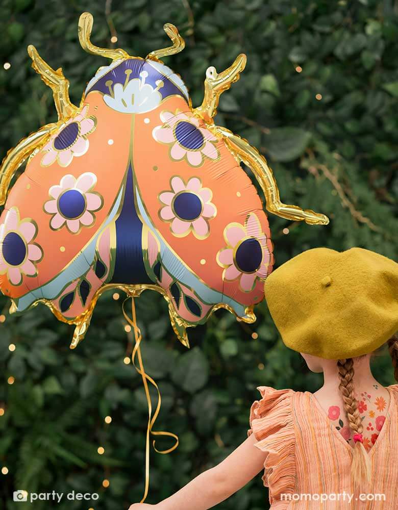 A little girl holding Momopartys's 34 x 30 inches ladybird shaped foil balloon by Party Deco. With coral color with small flower pattern with gold legs on it. She's dressed in her spring outfit with a mustard beret hat standing in a backyard with a wall filled of greenery.