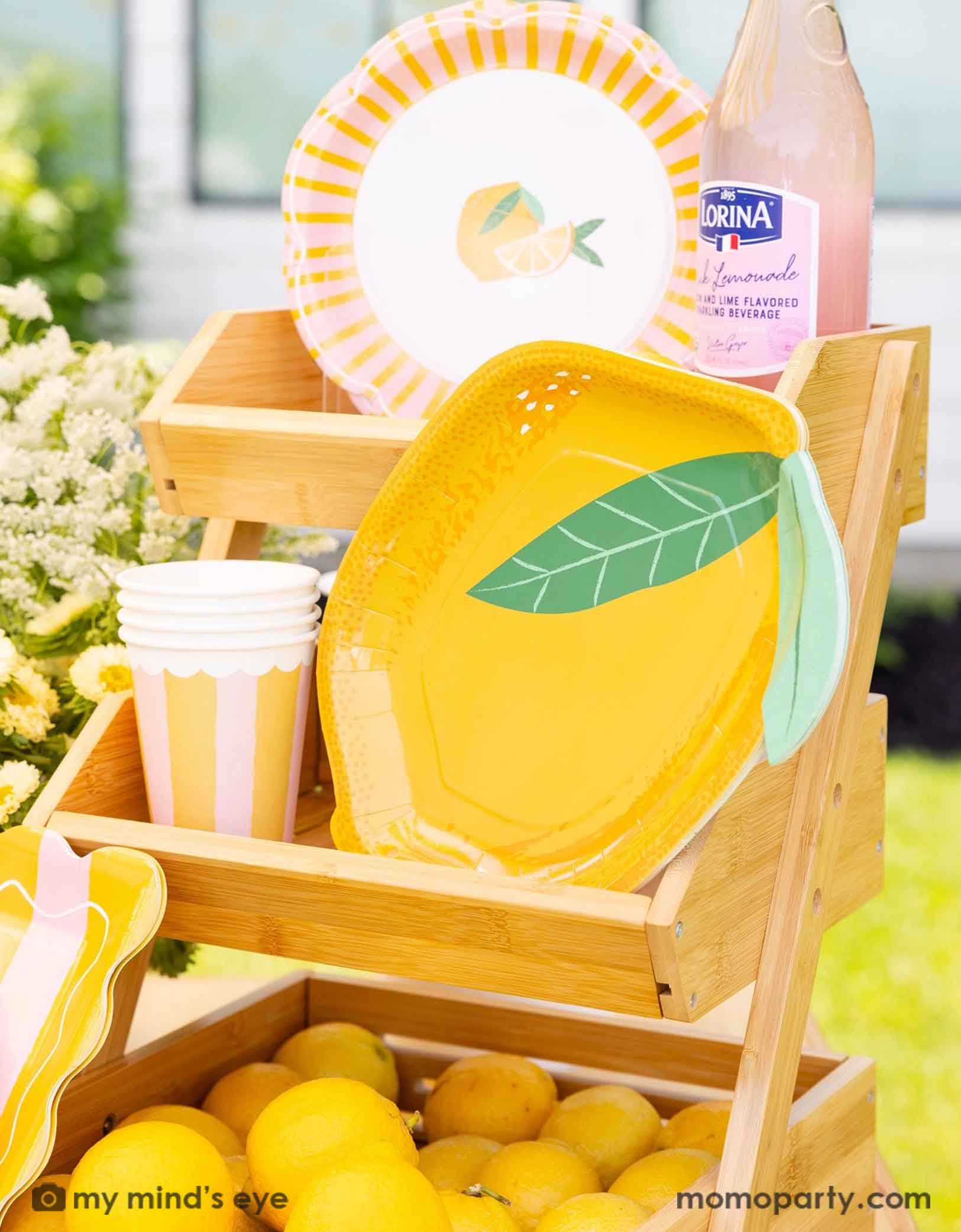 A 8.75" x 11.25" yellow lemon shaped plate with lemon themed party cups and side plates form Momopartys on a wooden stand. With a bottle of Lornia lemonade,  lemons and flowers in the background.