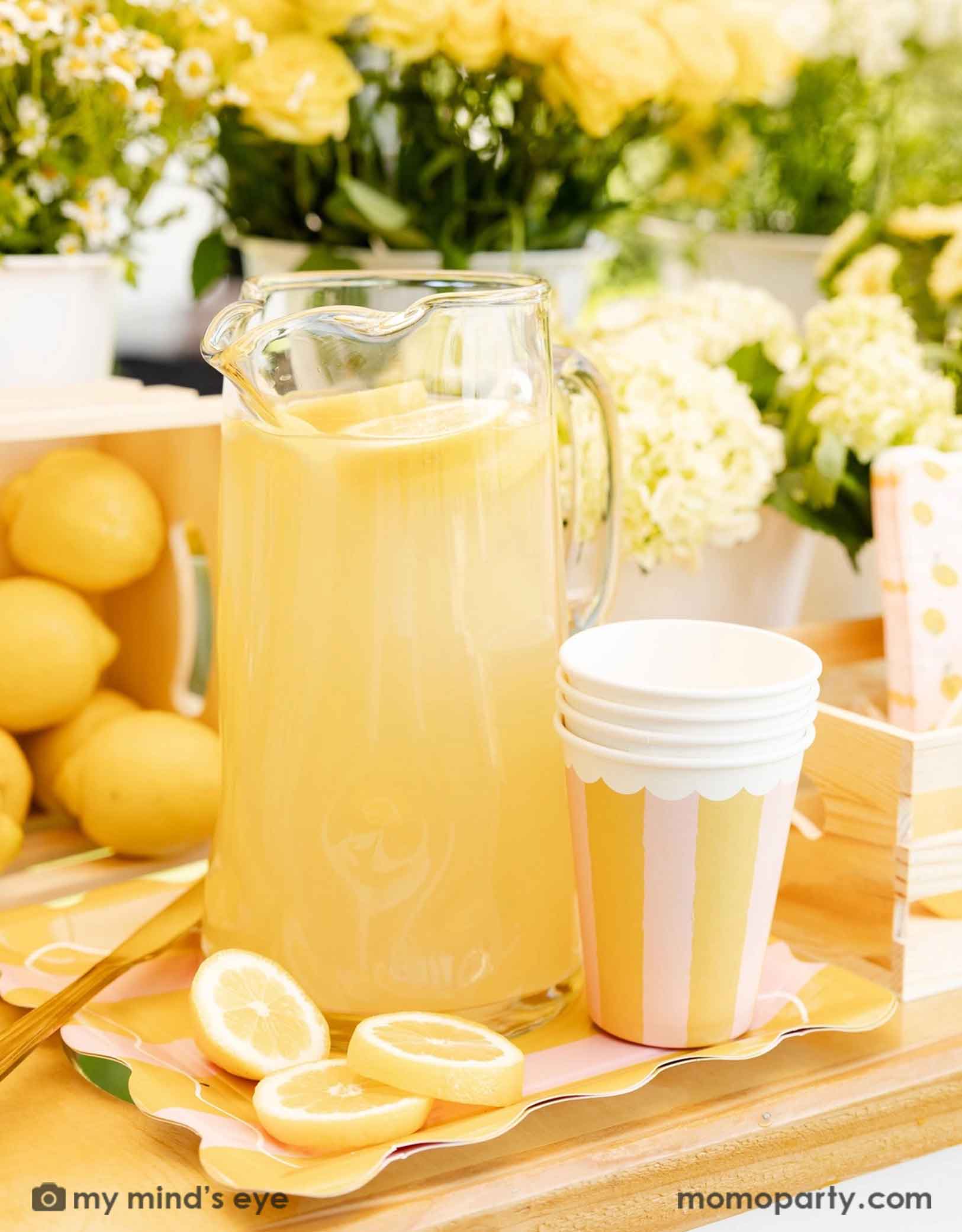 A glass pitcher of lemonade with lemons and Momopartys's Yellow & Pink Striped Party Cups next to it, on a wooden dessert cart with lemons and flowers in the background.