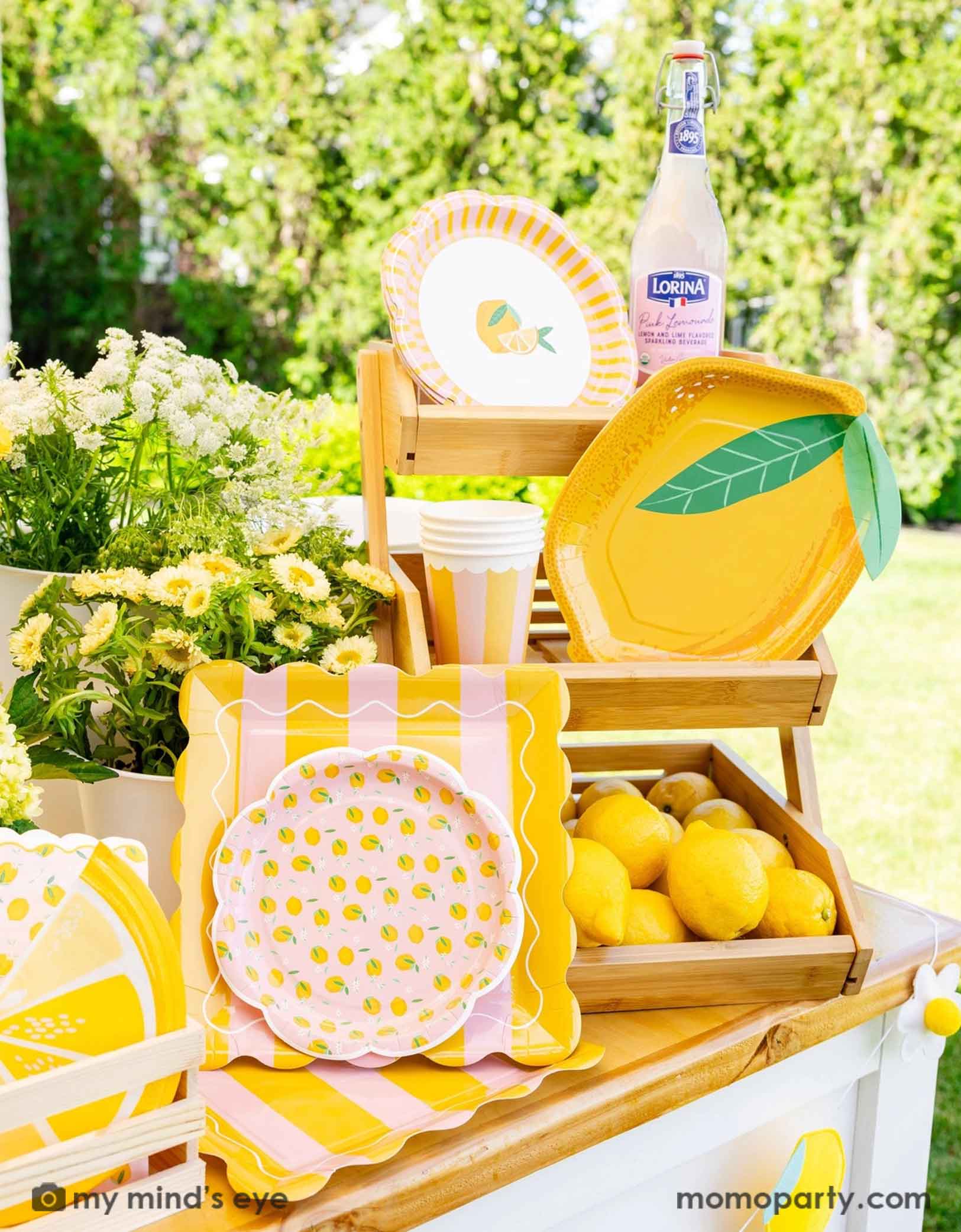An outdoor setting with lemon themed party supplies in yellow and pink from Momopartys including lemon shaped plates, napkins and party cups on a wooden stand. With lemons and flowers around, this cute little lemon stand is perfect for a summer gathering, be it a kid's lemon themed birthday, a baby shower or a bridal shower.