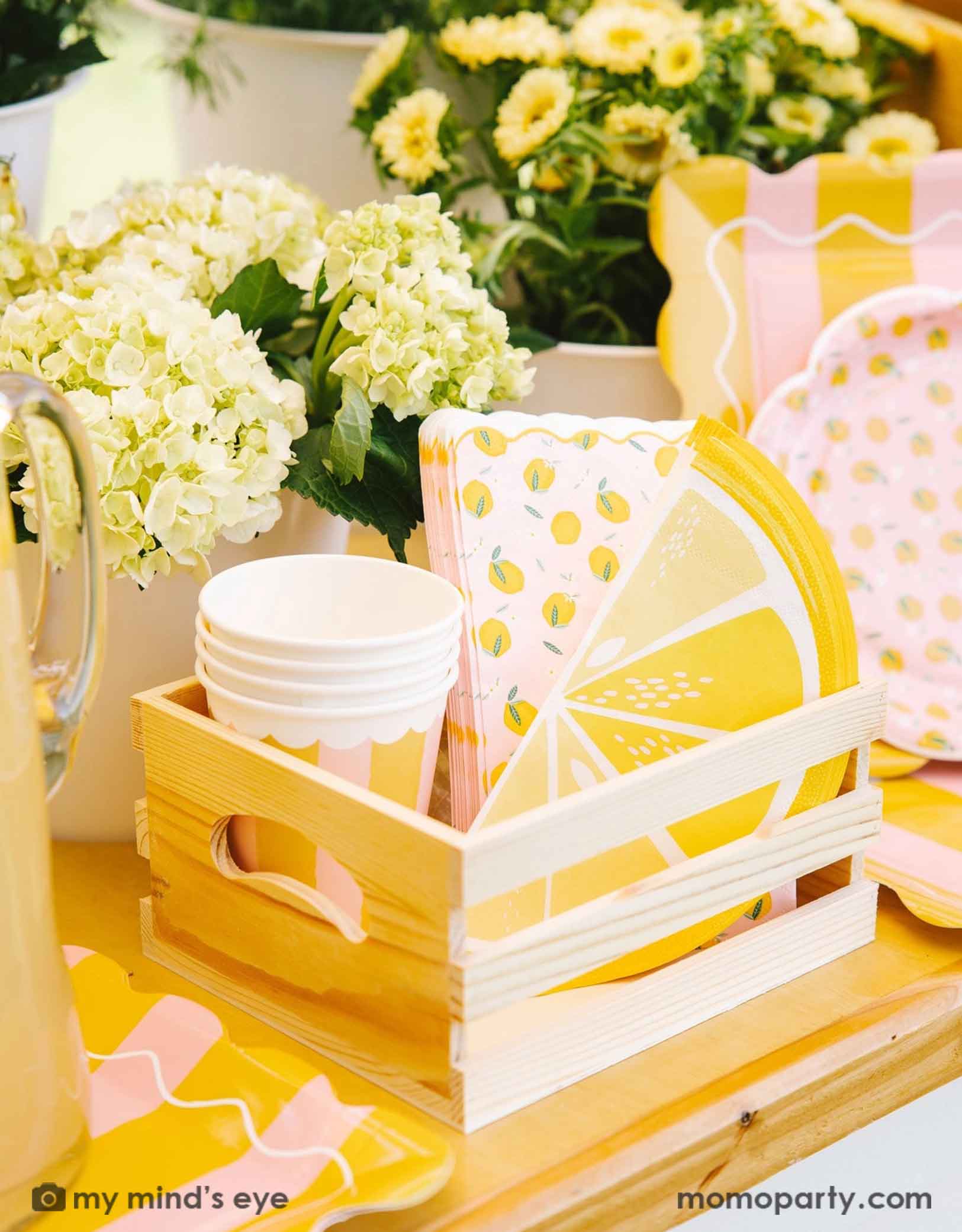 A cute wooden crate filled with party supplies with lemon design on plates, party cups and napkins from Momopartys, set against a beautiful floral background.