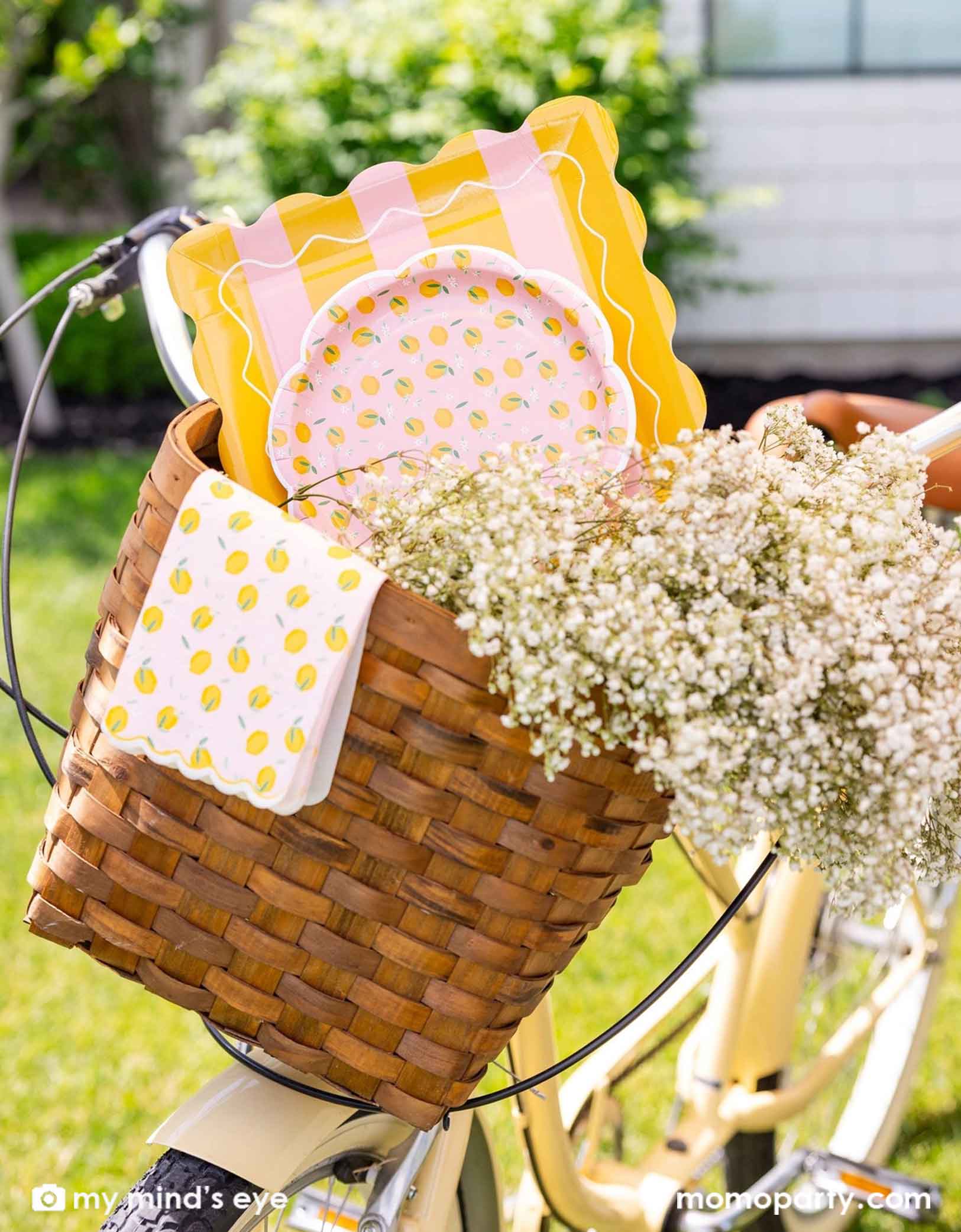 A cute bicycle basket with various of lemon themed party tablewares from Momopartys including the yellow striped square plates, lemon scattered round plates and lemon scattered dinner napkins. With flowers around, it makes an adorable scene.