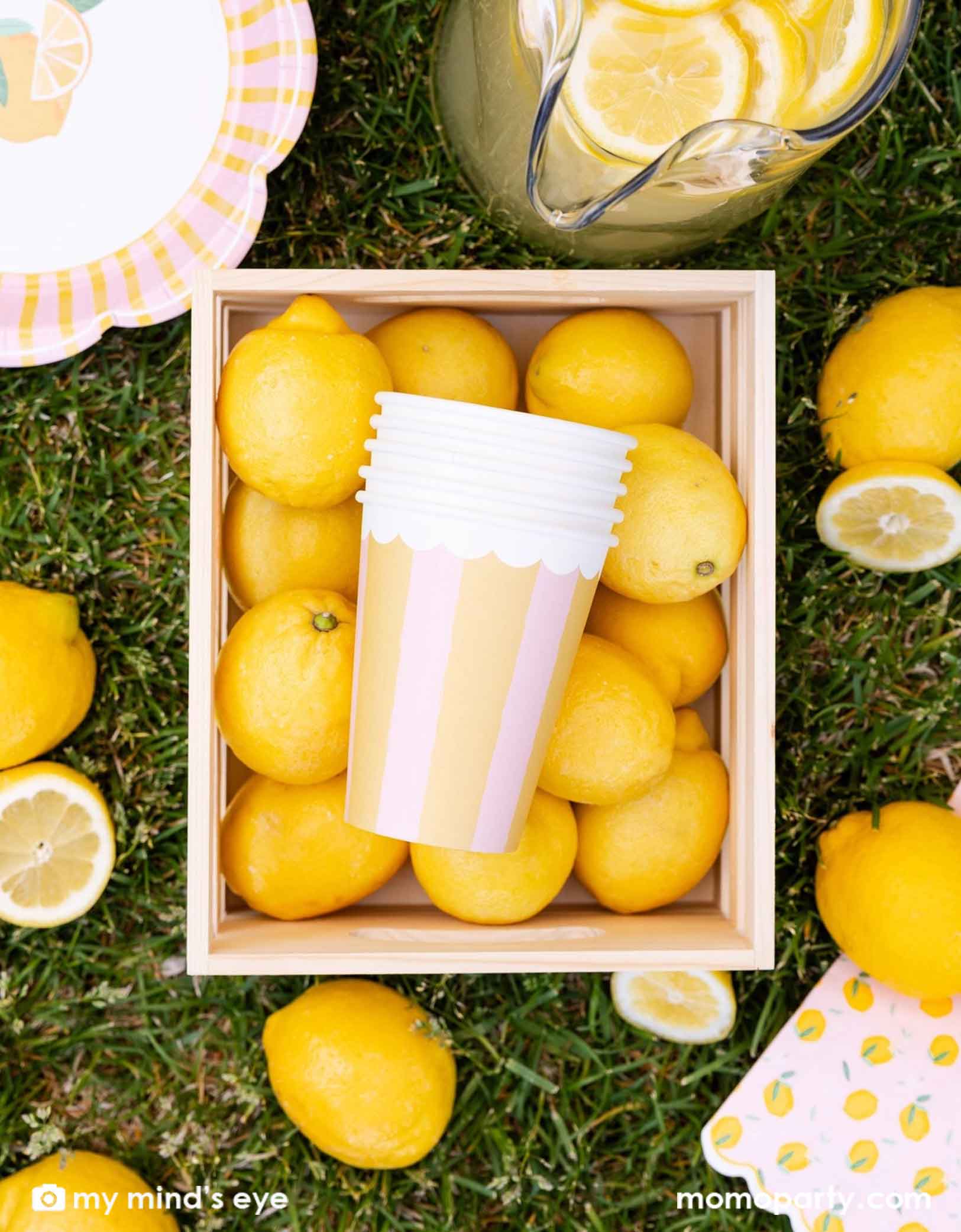 A wooden crate box of many lemons with Momopartys's Yellow & Pink Striped Party Cups in it, along with a pitcher of lemonade and lemon themed party plates and napkins on grass.