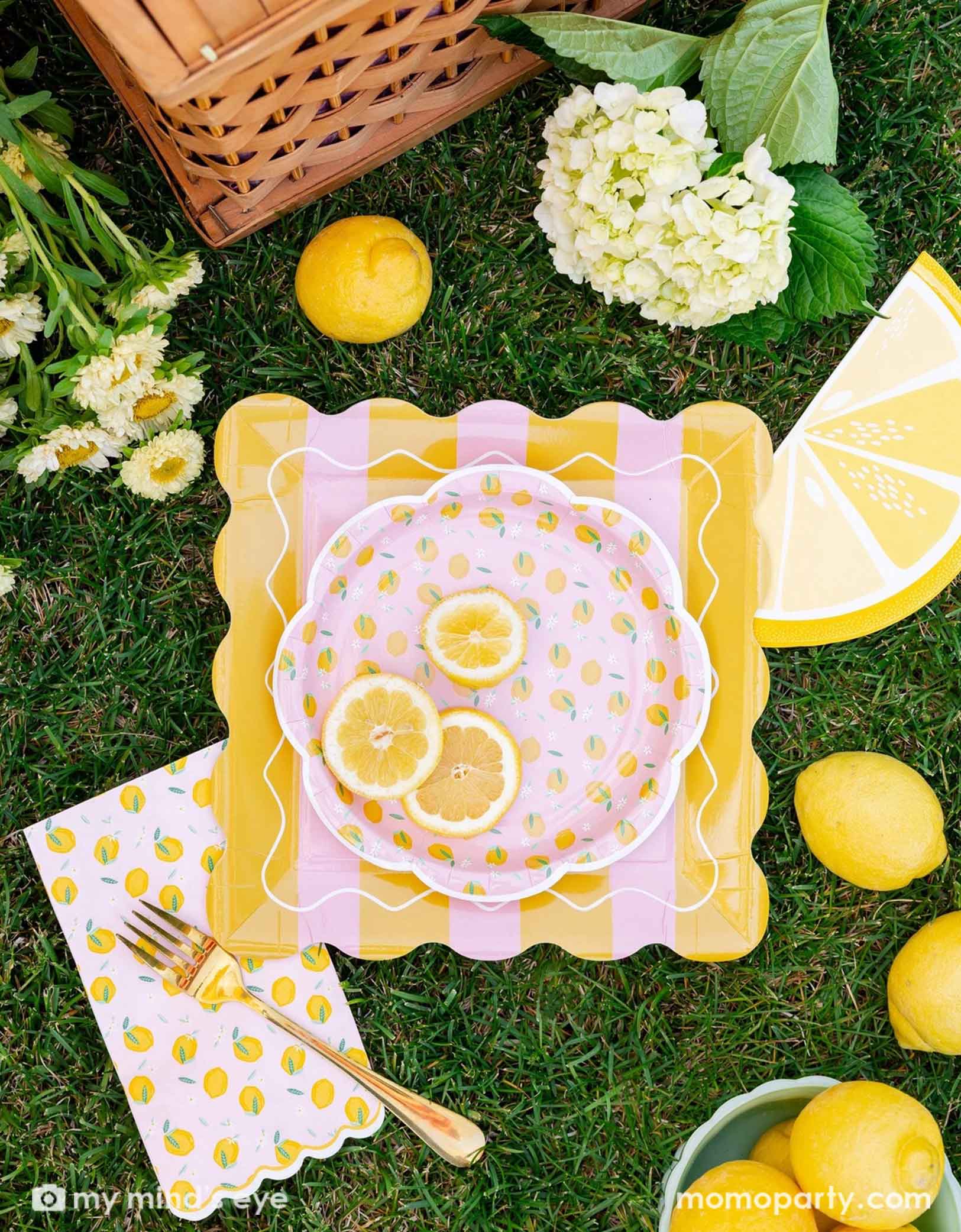 Decorative table setting featuring various lemon themed party tablewares from Momopartys including the 10" yellow and pink striped plate, lemon scattered round plate, lemon scattered dinner napkin and lemon slice shaped napkin. With flowers and lemons on a grassy surface.