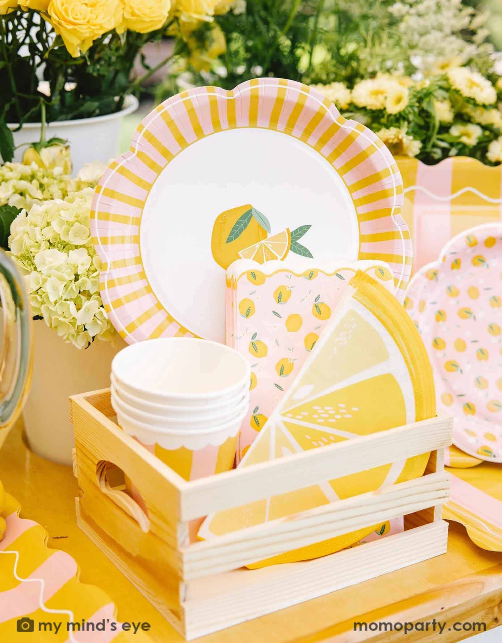 A cute wooden crate filled with party supplies with lemon design on plates, party cups and lemon sliced shaped napkins from Momopartys, set against a beautiful floral background.