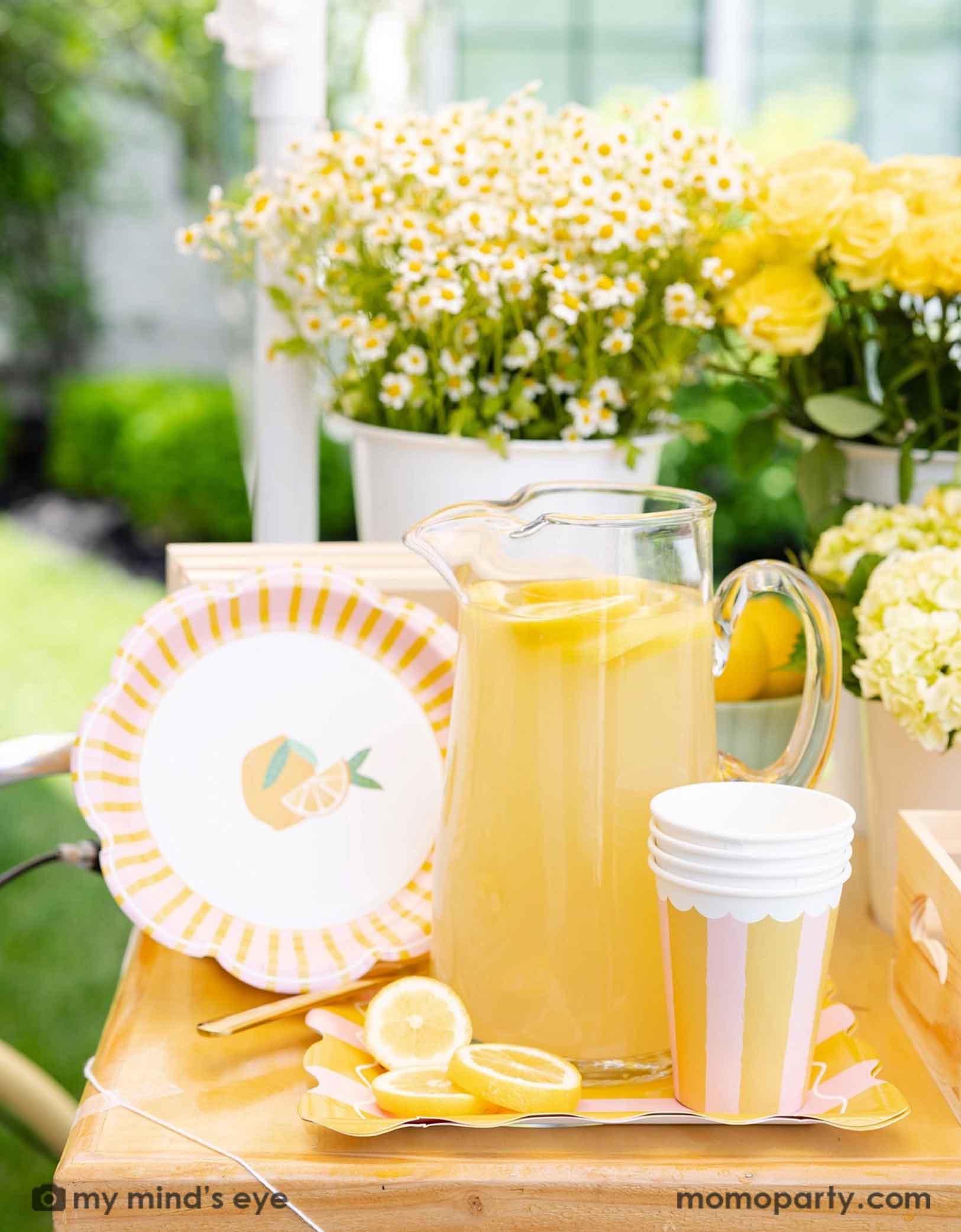 A glass pitcher of lemonade with lemons and Momopartys's Yellow & Pink Striped Party Cups and pink and yellow striped side plates next to it. With bouquets of flowers in yellow and white on a wooden dessert cart with lemons in the background. A beautiful dessert cart for a summer-y lemon themed celebration, be it a kid's birthday party, a baby shower, or a bridal shower.  