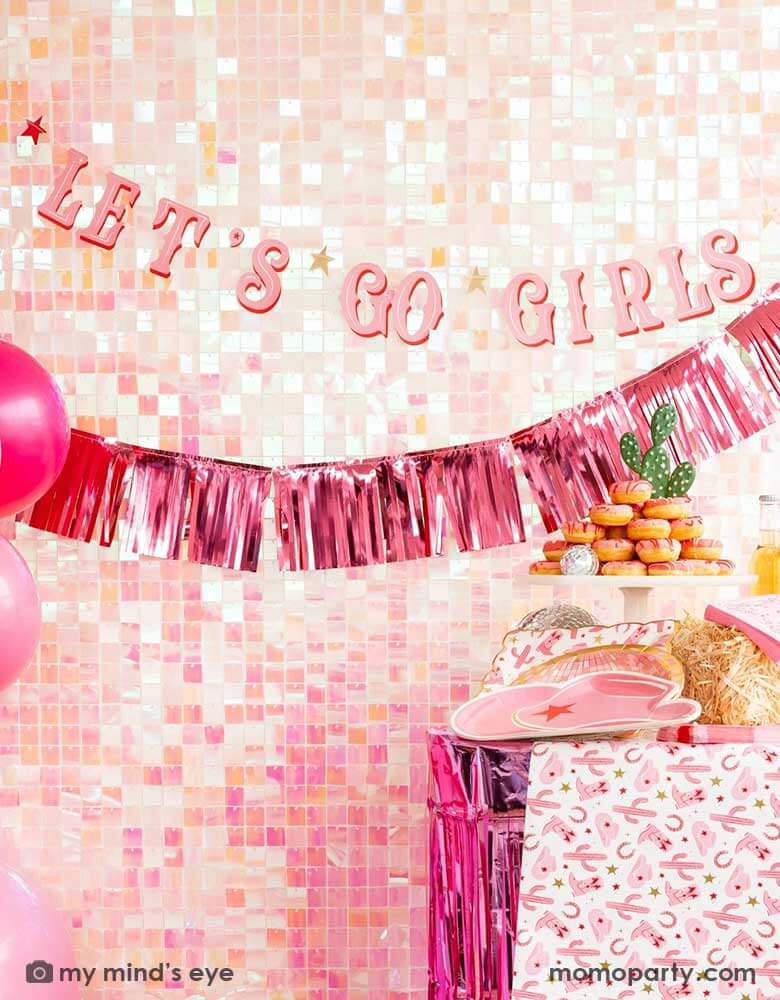  A cowgirl Western themed party table features pink cowgirl themed party supplies from Momopartys including pink cowgirl boot shaped napkins, cowgirl hat shaped plates, pink disco ball shaped plates, pink bandana small napkins, yeehaw party cups and cowgirl pattern paper plates and table runner by My Mind's Eye. In the back there's an iridescent backdrop adorned with Let's Go Girls pink foil fringe banners, along with a pink donut tower cake, making this a great inspo for a disco cowgirl party.