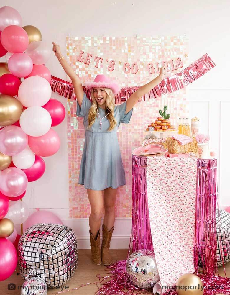 A young Caucasian lady in the Western inspired outfits and a pink cowgirl hat in a disco cowgirl bachelorette party decorated with cowgirl themed party supplies, decorations and balloons from Momopartys. She's raising her both hands by a table decorated with cowgirl party supplies including plates, napkins and a paper table runner. There's a donut cake, beverages and various treats on the table. Behind them is a iridescent wall adorned with "Let's Go Girls" pink foil fringe party banner from Momopartys.