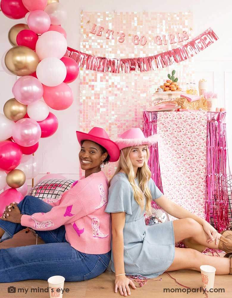 Two young ladies in Western inspired outfits and pink cowgirl hats in a disco cowgirl bachelorette party decorated with cowgirl themed party supplies, decorations and balloons from Momopartys. They're sitting back to back on the floor by a table decorated with cowgirl party supplies including plates, napkins and a paper table runner. There's a donut cake, beverages and various treats on the table. Behind them is a iridescent wall adorned with "Let's Go Girls" fringe party banner from Momopartys.