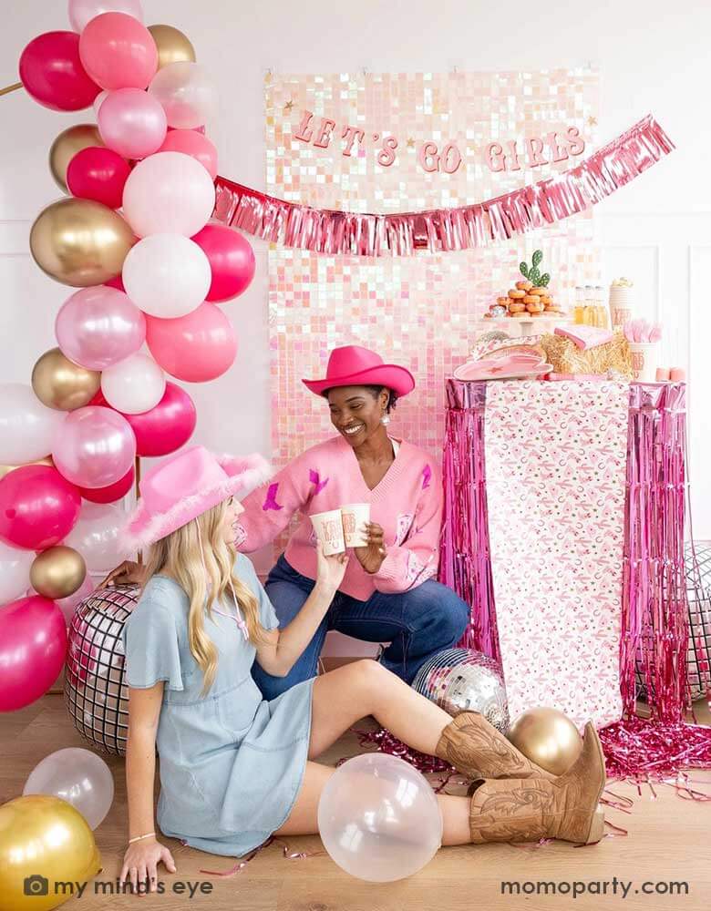 Two ladies with Western inspired outfits in a disco cowgirl bachelorette party decorated with cowgirl themed party supplies, decorations and balloons from Momopartys. They're raising their 12oz YEEHAW party cups by My Mind's Eye enjoying themselves in this festive celebration with donut cakes, beverages and various treats on the table decorated with cowgirl pattern table runner and pink foil fringe next to them. In the background there's a iridescent wall decorated with Let's Go Girls foil fringe banner.