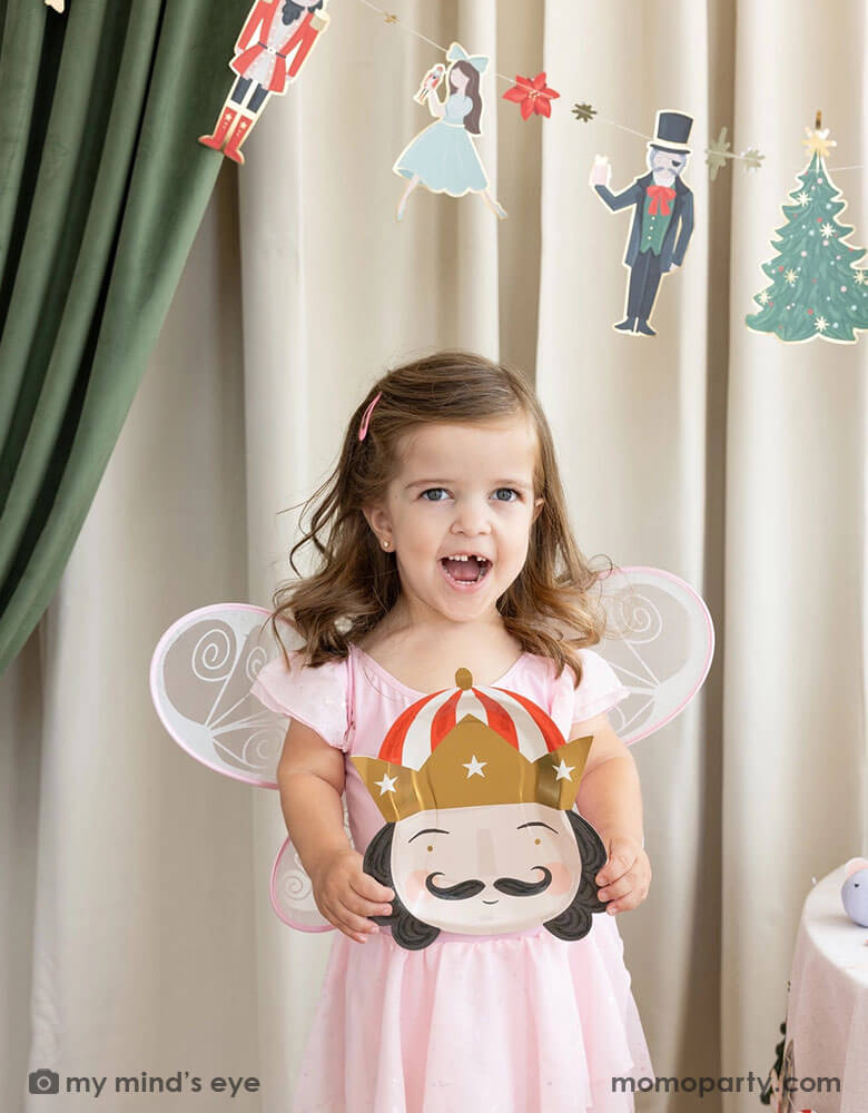 A little girl dressed up as Sugar Plum Fairy from Nutcracker's holding Momopartys's 10" Nutcracker head shaped party plate. In her back there's a stage curtain in satin green and beige which is decorated with Momopartys's Nutcracker garland with pom-poms which adds a festive touch to the Holiday decoration.