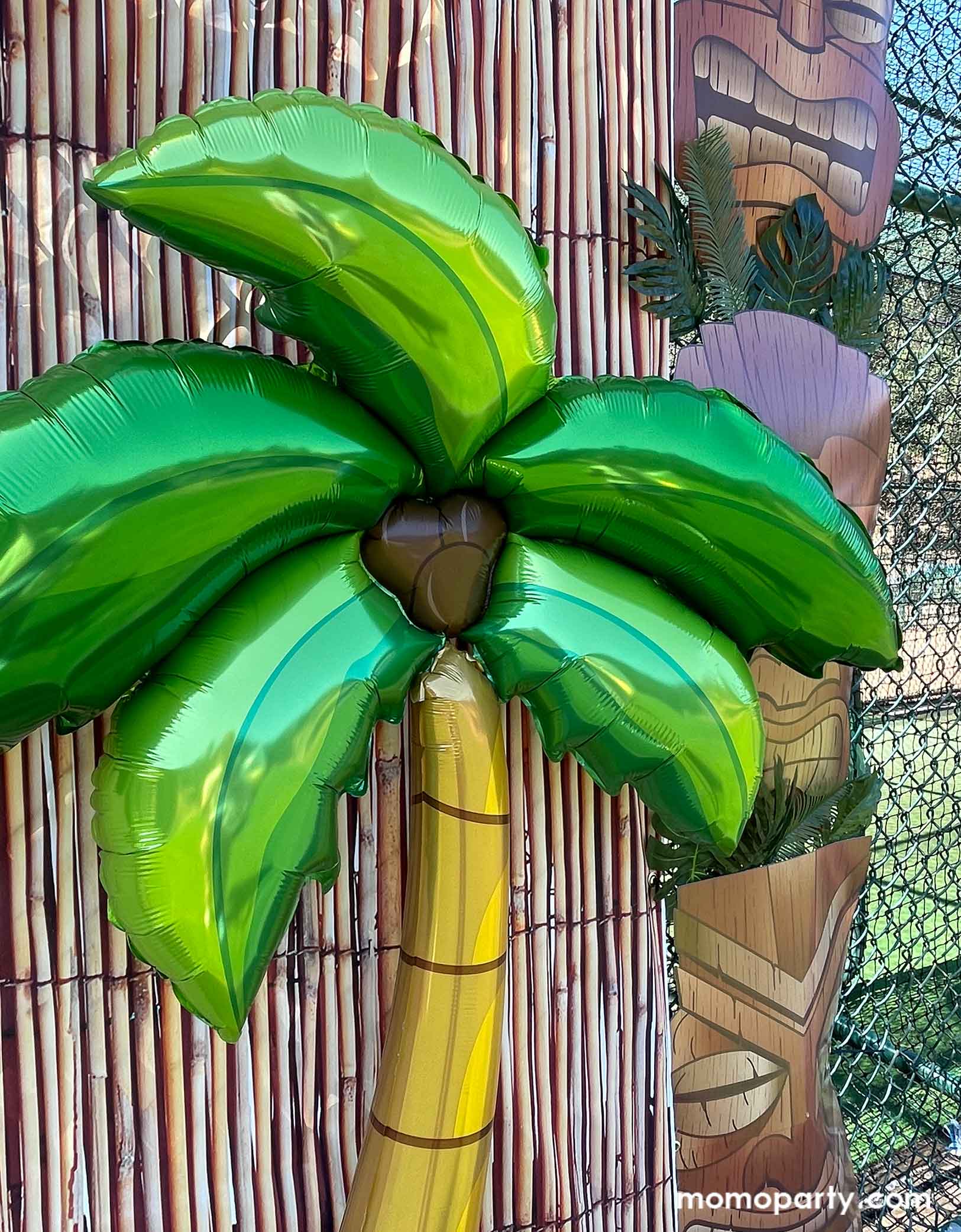 A close up shot of Momopartys's 65" Jumbo Palm Tree Shaped Foil Balloon in  in a luau, tropical, tiki themed party photo backdrop decoration featuring bamboo party backdrop, jumbo Tiki Mask Cardboard Cutouts adorned with palm leaves as two pillars. This palm tree shaped foil balloon is an easy and fun decoration to set a island scene for any tropical themed celebration, be it a birthday party, a baby shower, an anniversary celebration or a summer gathering.