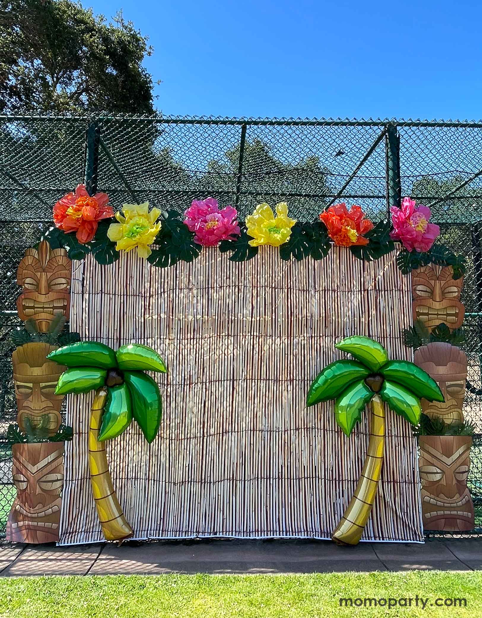 A luau, island, tiki, Hawaiian theme party photo backdrop decoration set up by Momopartys featuring bamboo party backdrop, jumbo Tiki Mask Cardboard Cutouts adorned with palm leaves as two pillars. On the top of the backdrop are some tissue paper Hawaiian hibiscus flowers in orange, yellow and pink colors along with palm tree leaf decorations. In the front are two 65" Jumbo Palm Tree Shaped Foil Balloons from momopartys.shop, making this a festive decoration idea for kid's island themed birthday celebration.