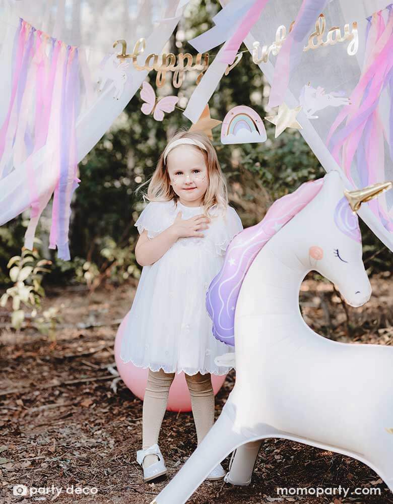 A girl in a white dress standing in an enchanting unicorn themed birthday party backdrop featuring Momopartys's 37.5" x 34" unicorn standing foil balloon around a gold Happy Birthday banner with pastel streamers hung in the front of a dreamy curtain, making this a whimsical scene for a girly's whimsical unicorn themed birthday party.