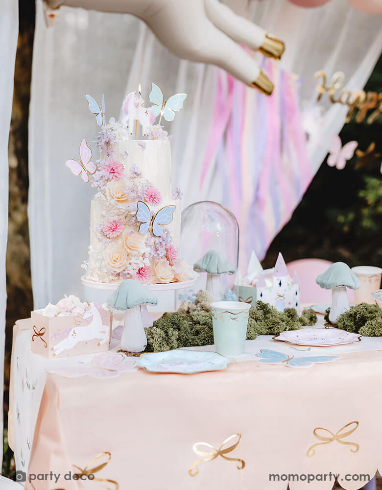 A closeup shot of a whimsical fairy butterfly themed birthday cake with beautiful floral buttercream, topped with Momopartys's pastel butterfly cake toppers. The table is covered with Momopartys's pink bow paper tablecloth with gold foil accent. On the table there are pastel colored tableware including paper plates, party cups, treat boxes and party favor bags. In the middle there's faux grass and mushroom decoration, making this an enchanting set up for girl's magical birthday celebration.