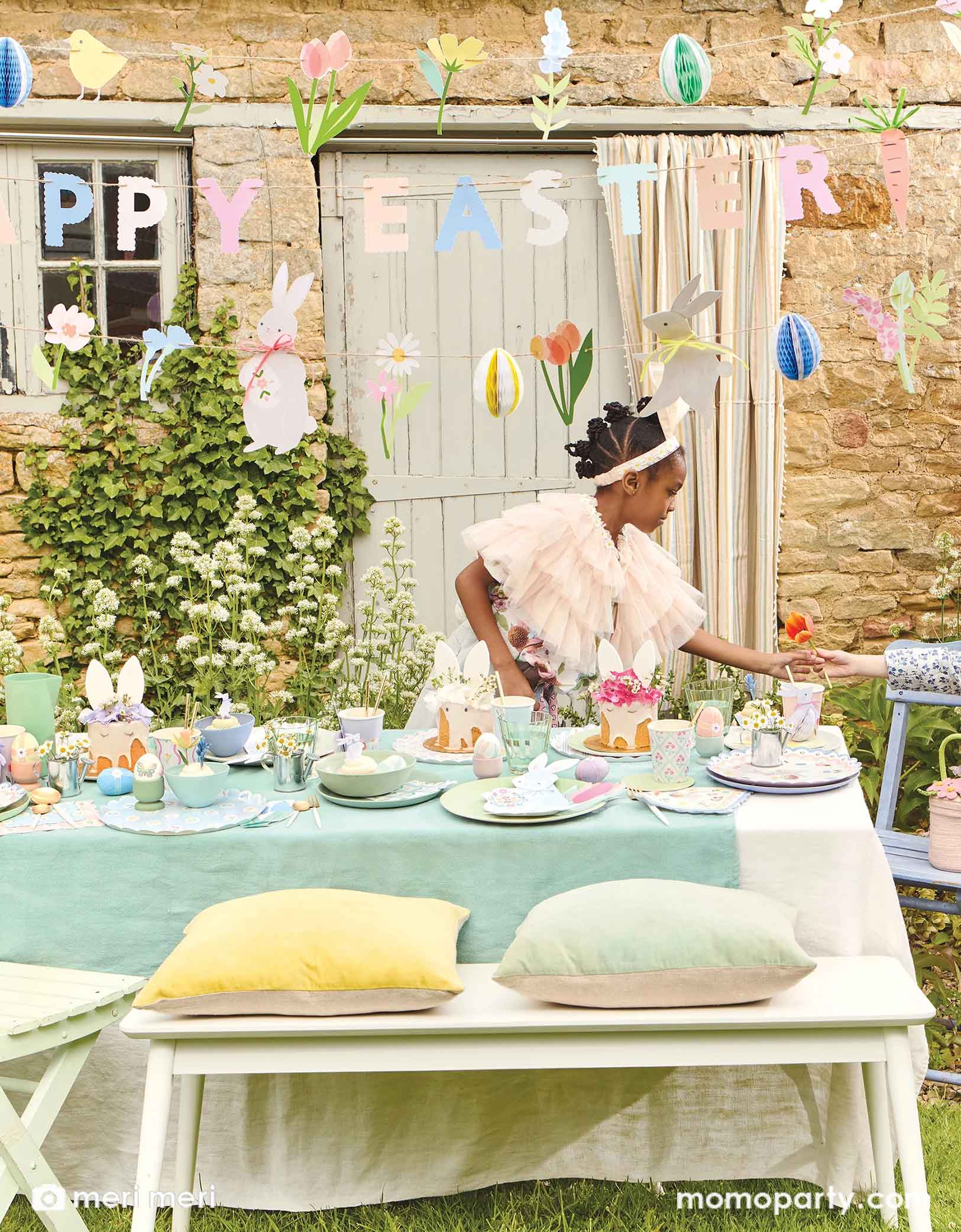 A girl wearing a bunny headband stands next to an Easter party table with Meri Meri's Easter party supplies including the Easter bunny shaped plates, gingham checkered Easter cupcake kit, Easter bunny napkins, with pastel aqua blue tablecloth, with spring tulip flowers and Easter eggs and treats on the table. Above the table hung a Happy Easter party garland featuring floral pennants and egg honeycombs, making this a darling setup to celebrate Easter with your kids.