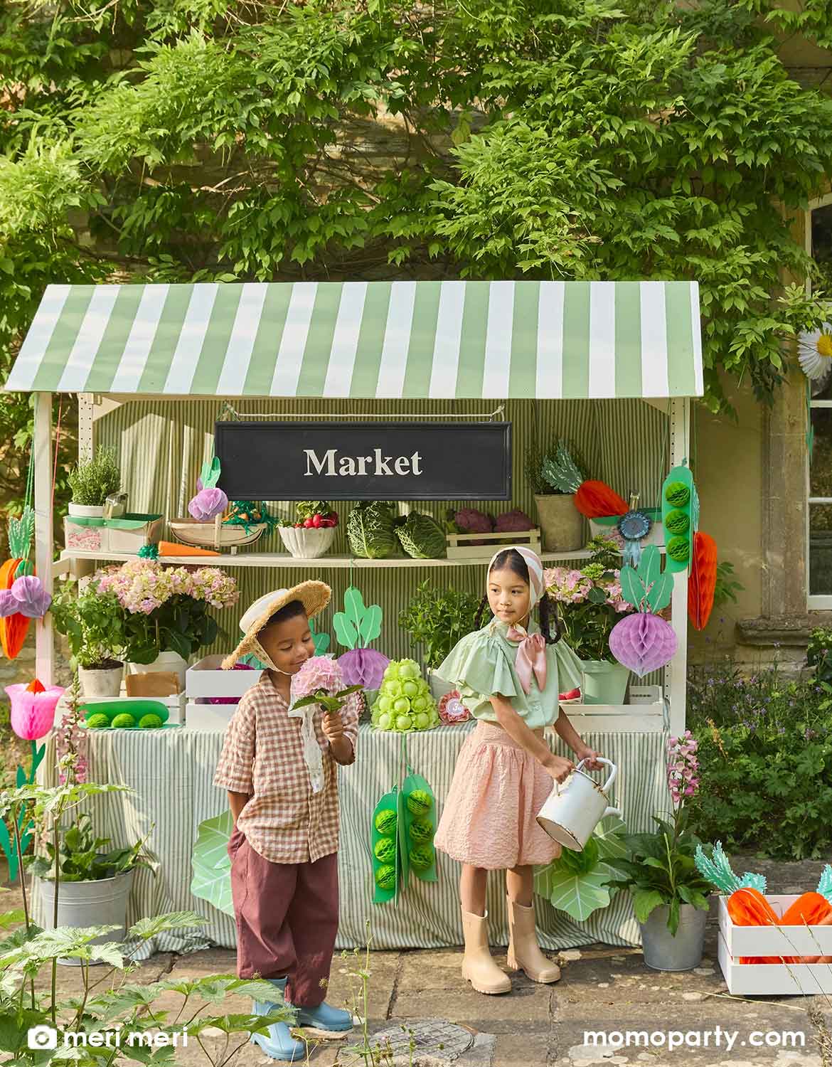 Two children playing at a pretend farmer's market stand with a light green striped canopy that's decorated with Momopartys's giant vegetable honeycomb decorations hanging under the canopy including carrots, beets, lettuces and green pees by Meri Meri. With flowers, plants, produces and various props on the stand. An adorable set up for kid's farmer's market inspired birthday party.