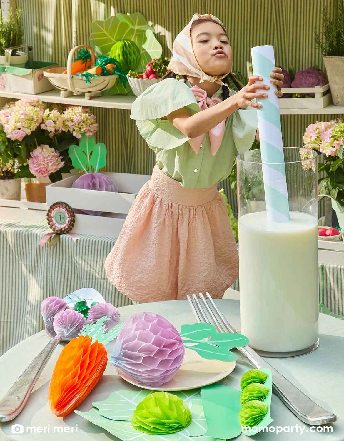 A girl in a cottage inspired outfits using a giant straw to drink a giant cup of milk at a garden themed party table with Momopartys's vegetable honeycomb decorations including carrots, beets, lettuces and green pees by Meri Meri. Beyond her is a pretend farmer's market stand with a light green striped canopy, with plants, produces and various props on the stand. Making this a whimsical inspiration for a kid's farmer's market themed birthday party.