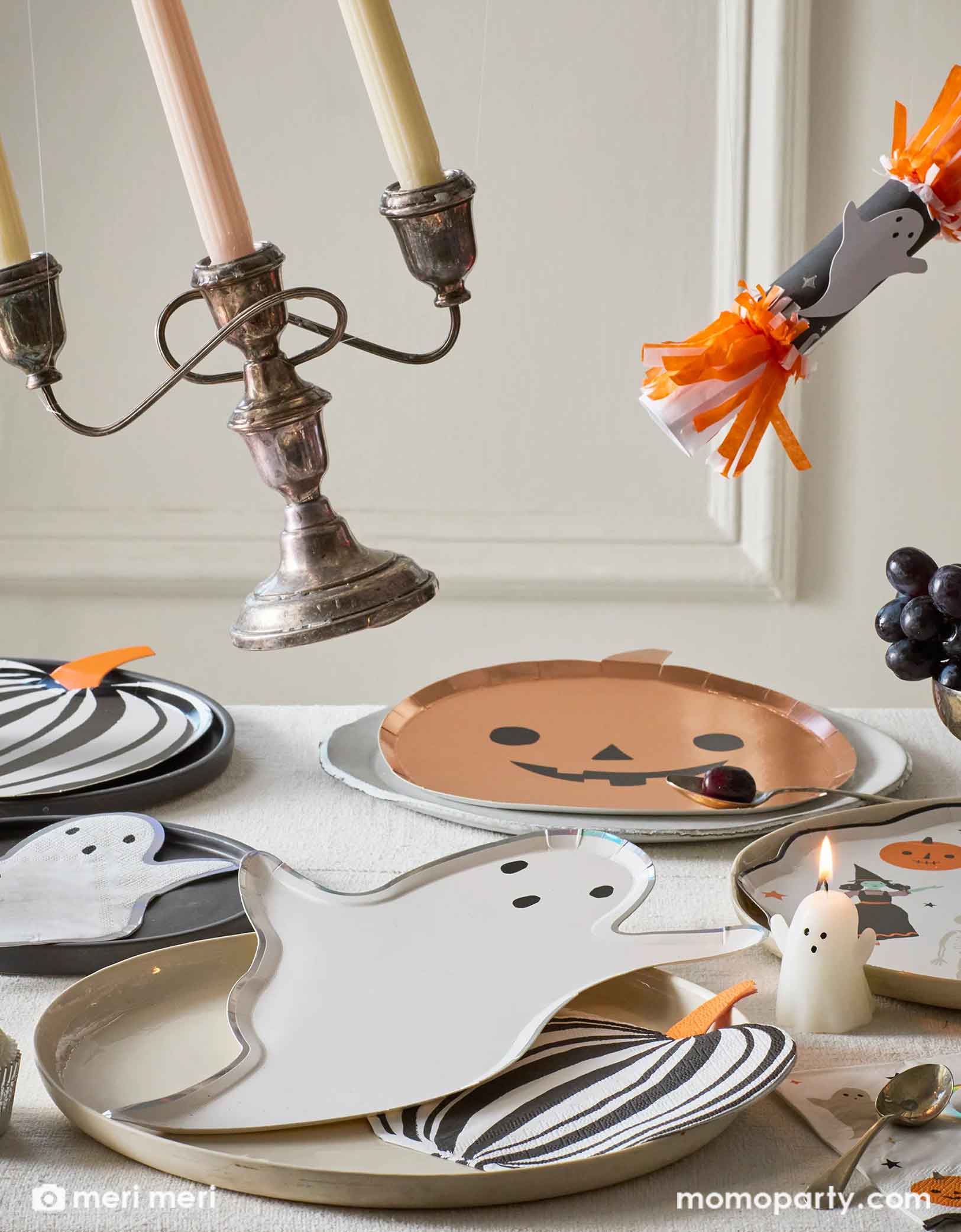 A Halloween-themed table setting with an array of Momopartys's Halloween party supplies including ghost and pumpkin shaped plates and matching napkins, black striped pumpkin plates and napkins by Meri Meri. With ghost candles, Halloween ghost party crackers and decorative items flying off on the table, this creates a fun scene for a kid's friendly Halloween celebration in this spooky season!