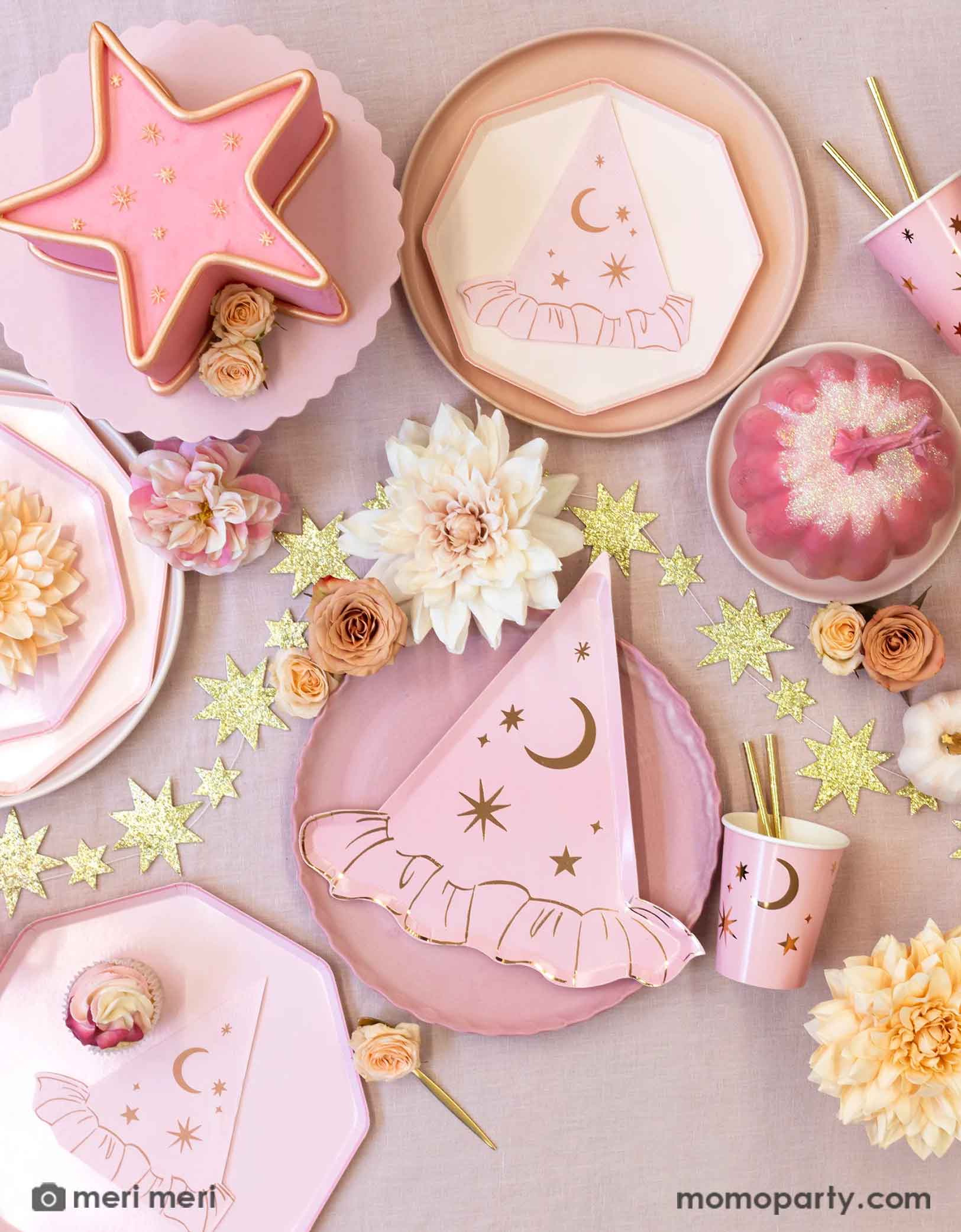 An overhead shot of a pink Halloween party table featuring Momopartys's pink witch themed party supplies with star and moon designs including the pink dinner plates with witch hat shaped plates, napkins and pink party cups with gold foil straws. Around the party tablewares are some dahlia flowers in blush and pale pink, a pink star shaped cake and the gold star glittered garland spread across the table covered with pink tablecloth, making this a chic party look for a non-spooky Halloween celebration!