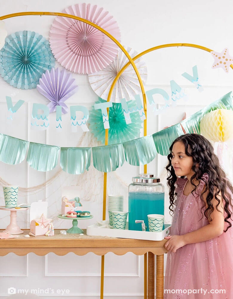 A girl in a pink tutu dress by a white wall decorated with mermaid party decorations including Momopartys's under the sea paper fans in pastel colors of aqua, pink, lilac, teal, cream and peach. Next to the paper fans hung a fishing net draping over a golden arch structure adorn with mermaid Happy Birthday teal foil banner by My Mind's Eye. The girl is standing next to a table decorated with mermaid supplies including mermaid tail party cups, under the sea treat boxes filled with treats and sugar cookies.