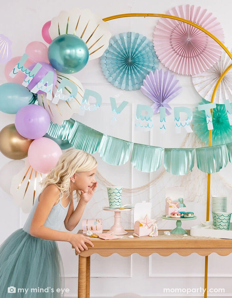 A girl in a teal tutu dress by a white wall decorated with mermaid party decorations including Momopartys's under the sea paper fans in pastel colors of aqua, pink, lilac, teal, cream and peach. Next to the paper fans hung a fishing net draping over a golden arch structure adorned with honeycomb jelly fish decorations by My Mind's Eye. She's whispering next to a table decorated with mermaid supplies including mermaid tail party cups, under the sea treat boxes filled with treats and sugar cookies.