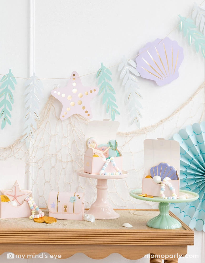 A mermaid party decoration set up on the wall features Momopartys's under the sea paper fan set in pastel colors aand the jumbo mermaid garland featuring seagrass, sea stars and sea shells in dreamy pastel colors by My Mind's Eye, underneath hung a fishing net draping over the party table which has multiple treasure box shaped treat boxes filled with treats and mermaid themed sugar cookies, making this an perfect inspo for an enchanting kid's mermaid themed birthday party set up.