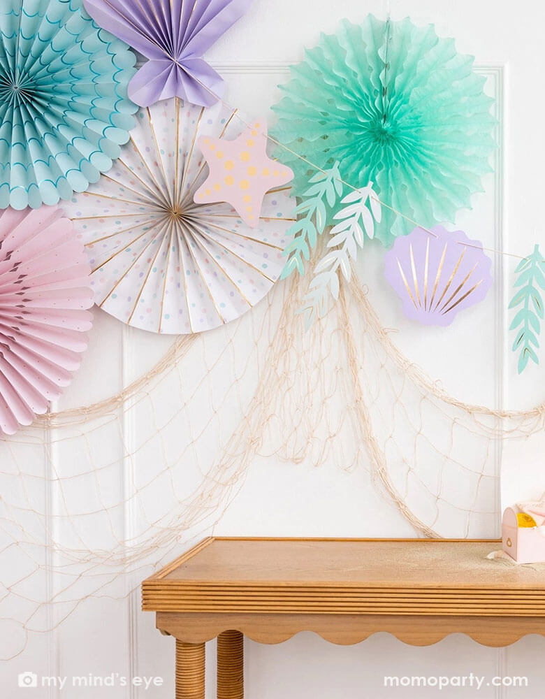 A mermaid party decoration set up on the wall features Momopartys's under the sea paper fan set which includes a set of 6 paper fans 6 fans of size 14", 8.5", 8", 7", and 5.5" in pastel colors of aqua, pink, lilac, teal, cream and peach. Next to the paper fans hung the jumbo mermaid garland by My Mind's Eye in the matching colors, underneath hung a fishing net draping over the party table, making this an perfect inspo for an enchanting kid's mermaid themed birthday party set up.