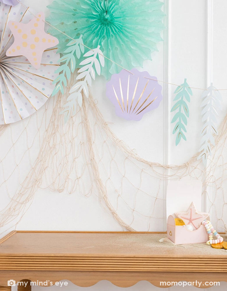 A mermaid party decoration set up on the wall features Momopartys's under the sea paper fan set which includes a set of 6 paper fans 6 fans of size 14", 8.5", 8", 7", and 5.5" in pastel colors of aqua, pink, lilac, teal, cream and peach. Next to the paper fans hung the jumbo mermaid garland by My Mind's Eye in the matching colors, underneath hung a fishing net draping over the party table, making this an perfect inspo for an enchanting kid's mermaid themed birthday party set up.
