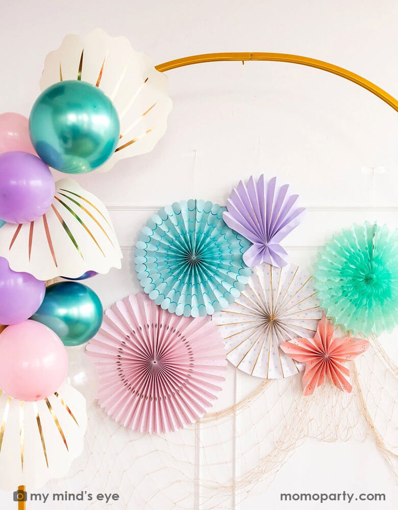 An elegant white wall decorated with mermaid inspired party decorations from Momopartys including under the sea paper fan set in pastel colors colors of aqua, pink, lilac, teal, cream and peach, along with mermaid themed party balloons in coordinating colors with My Mind's Eye's large cream shell shaped paper decorations, with a fishing net hung underneath, this makes a perfect backdrop for an enchanting kid's under the sea, mermaid birthday party.