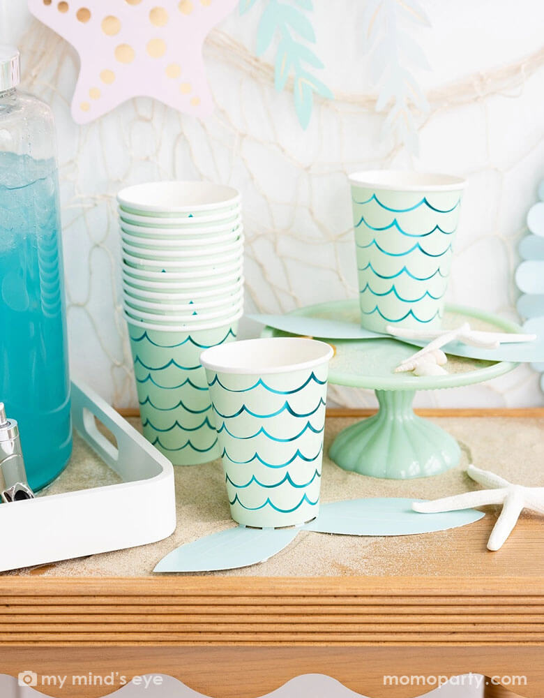 A mermaid party table features Momopartys's mermaid tail paper cups by My Mind's Eye. Next to the paper cups is a water dispenser filled with aqua colored drink. The table is decorated with shells, sea star and white sand decorations. In the back there's a fishing net hung on the wall which is adorned with under the sea paper party pennants in soft pastel colors, making this a perfect inspo for a kid's mermaid, under the sea party decorations. 