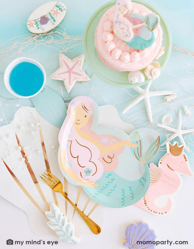 An enchanting mermaid party tablescape features Momopartys's mermaid shaped plate, pink seahorse shaped napkin, and mermaid tail party cup on a cream seashell shaped placemat by My Mind's Eye. Around these under the sea inspired tableware, there are mermaid themed sugar cookies and a small pink cake, along with some sea star decorations and fishing net, this makes a perfect inspo for kid's magical mermaid party ideas.