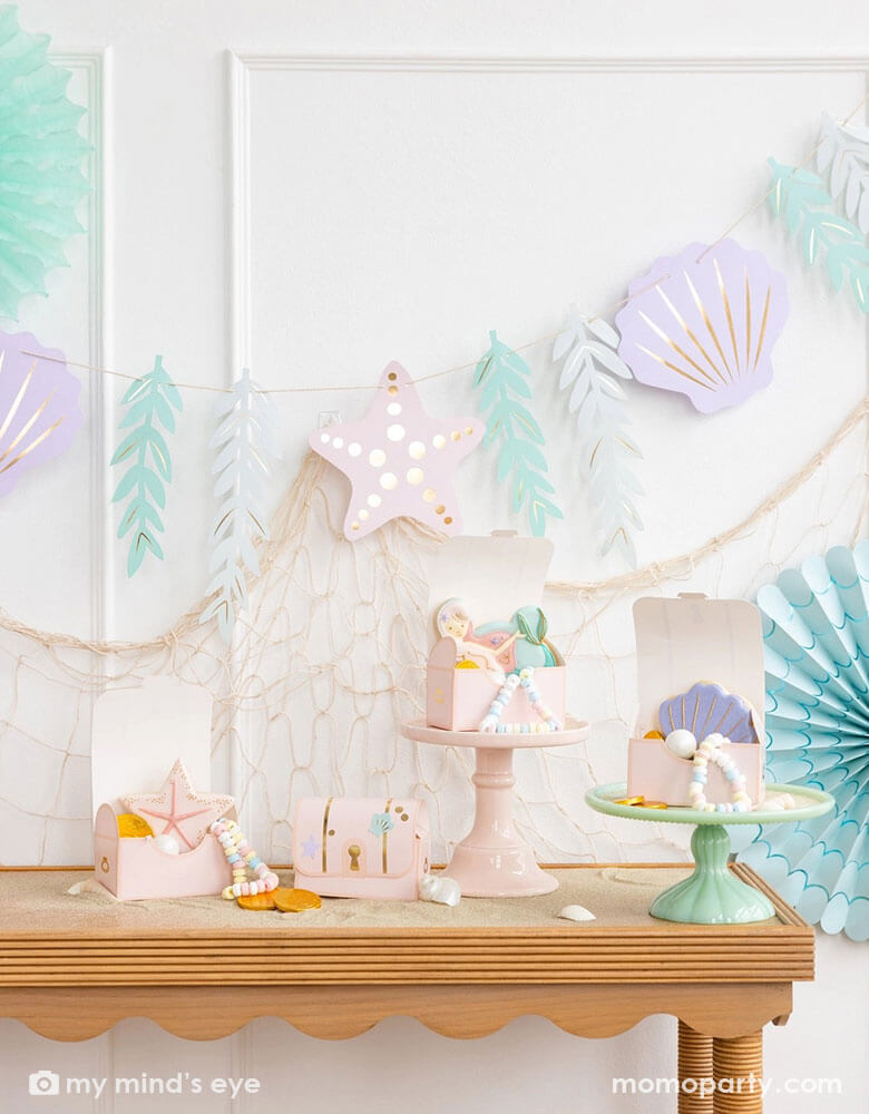 A mermaid party decoration set up on the wall features Momopartys's under the sea paper fan set in pastel colors aand the jumbo mermaid garland featuring seagrass, sea stars and sea shells in dreamy pastel colors by My Mind's Eye, underneath hung a fishing net draping over the party table which has multiple treasure box shaped treat boxes filled with treats and mermaid themed sugar cookies, making this an perfect inspo for an enchanting kid's mermaid themed birthday party set up.