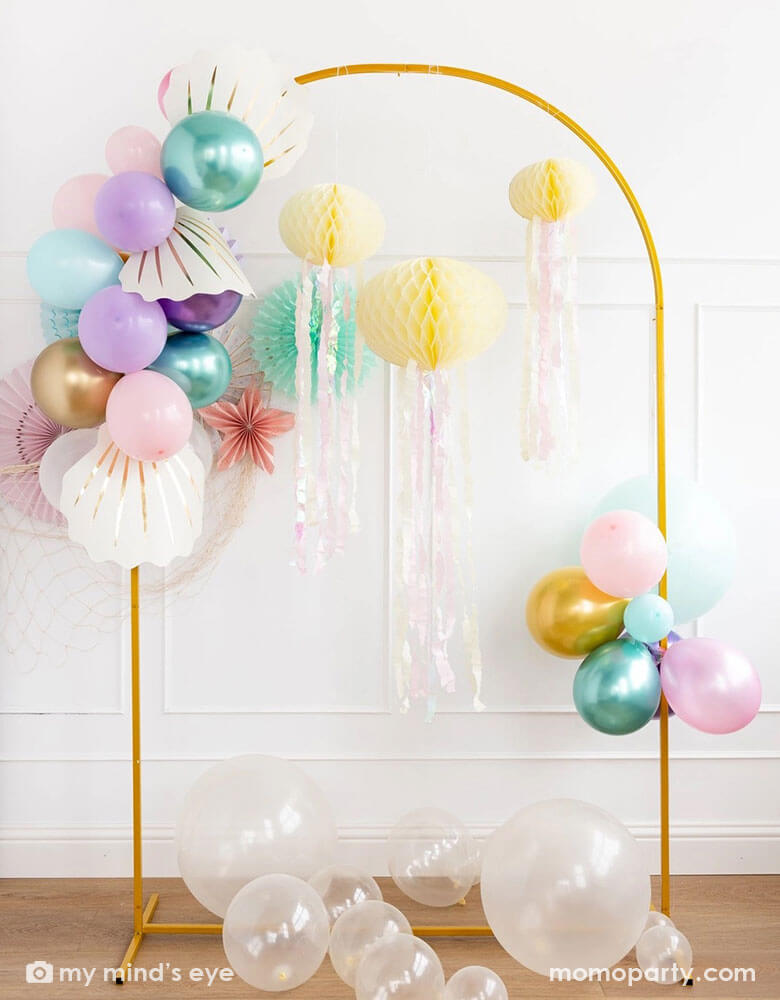 A white wall decorated with mermaid party decorations including Momopartys's under the sea paper fans in pastel colors. Next to the paper fans hung a fishing net draping over a golden arch structure adorned with honeycomb jelly fish decorations by My Mind's Eye. On the left side there is a mermaid inspired balloon garland adorned with shell shaped paper decorations in cream. On the floor there are some clear latex balloons as bubbles, making this an inspo for an enchanting kid's mermaid party set up.