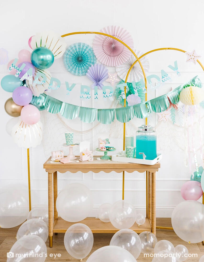 An enchanting mermaid party set up features Momopartys's mermaid decorations and and supplies by My Mind's Eye including  including the Under the Sea paper fan set in pastel colors of light pink, aqua, teal, lilac and cream, Mermaid Happy Birthday Banner Set with aqua foil fringe banner and shell shaped paper decorations on the wall. On the table there are various mermaid themed tableware including cups, napkins, plates and treat boxes. On the floor are clear latex balloons creating a whimsy bubble scene.