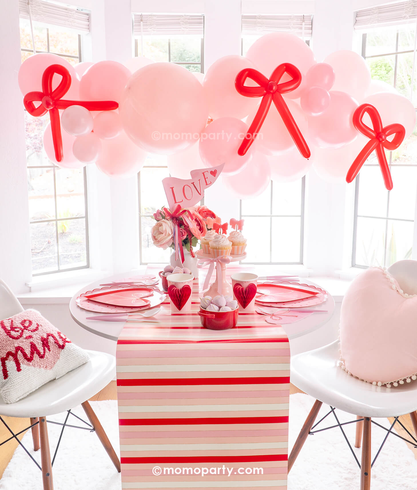 A beautiful party set up next to a big, bright window featuring Momopartys's Bow-tiful Valentine's Day Party Box for 8 guests. Featuring a pink balloon garland with red bow balloons on it, heart shaped tableware including plates, napkins and party cups, and a beautiful flower bouquet with a pink "LOVE" party pennant, this adorable party box in classic Valentine's colors of red and pink is perfect for your Valentine's Day party or Galentine's Day celebration.
