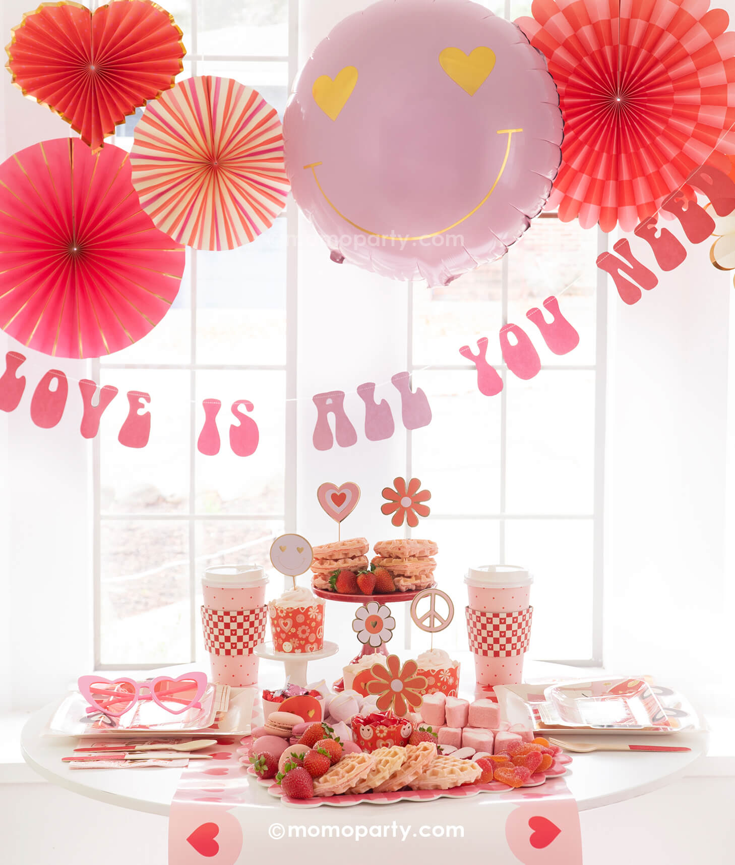 A Valentine's Day celebration featuring Momopartys's Groovy Valentine's Day Party box set up with pink and red paper fans in heart shape and a "love is all you need" banner in retro type hung below the paper fans. On the table you can see a sweet board featuring treats and snacks in red and pink colors including heart shaped waffles, strawberries, macarons, marshmallows etc. Next to the desert board featured retro inspired Valentine's plates, party cups and napkins,  a perfect inspo for Vday celebration!