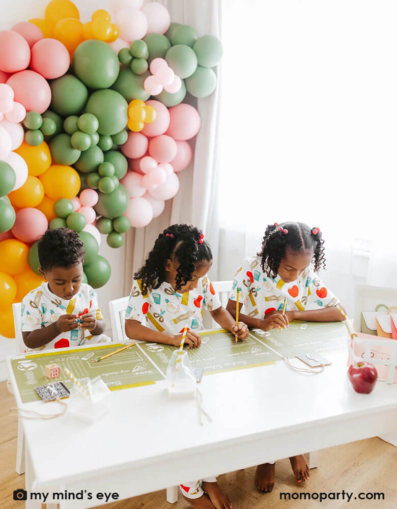 A kid's back to school first day of school celebration set up featuring a colorful balloon wall in the back with three school aged kids playing word games on Momopartys's back to school placemats by My Mind'd Eye. Each of the kid is wearing cute back to school pajamas, makes it a great idea to celebrate the first day of school with your kids.