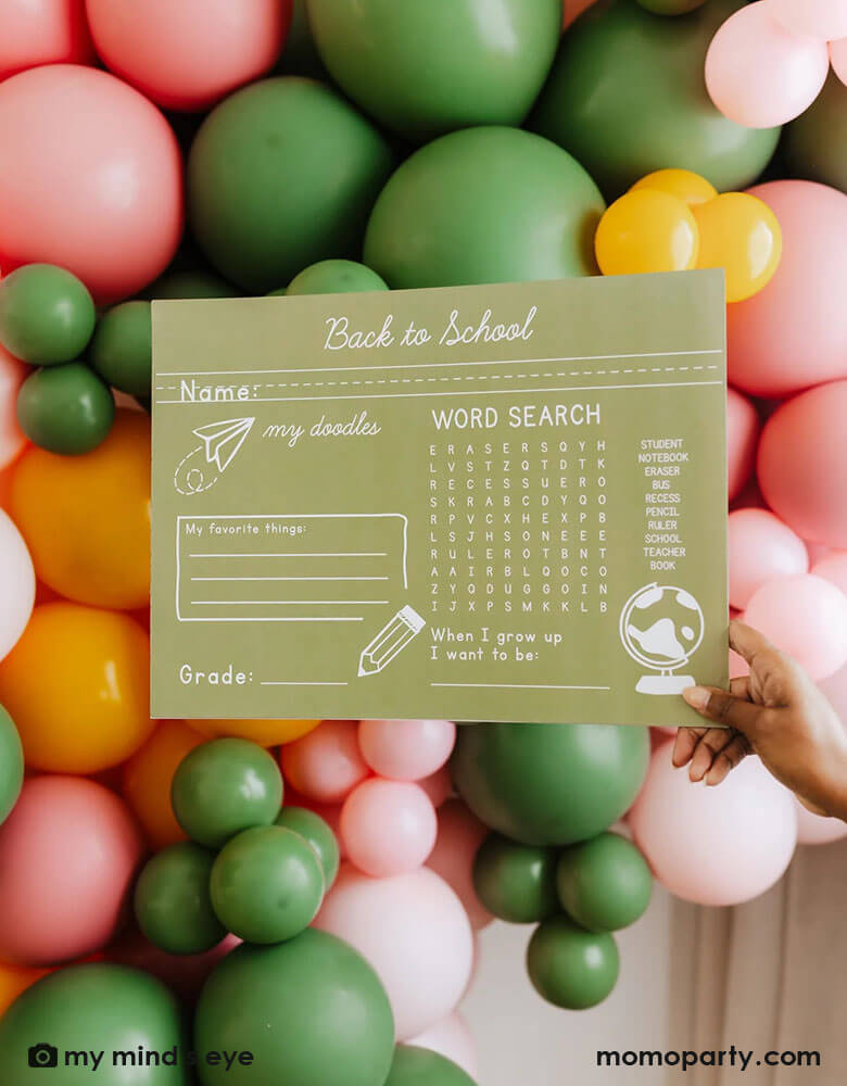 A mom holding Momopartys's 15.5 x 11.5 back to school placemat in front of a colorful balloon wall to celebrate kid's first day of school.