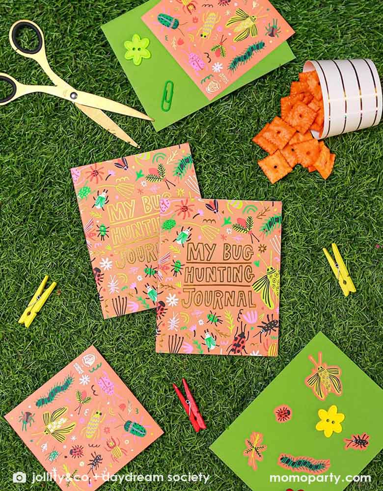 Two kid's bug hunting journals and Momopartys's backyard bug sticker sheets with some crafting tools laying on a lawn, with a foodcup filled with cheez-it crackers, makes it a cool party activity for a kid's bug themed birthday celebration or playdate.